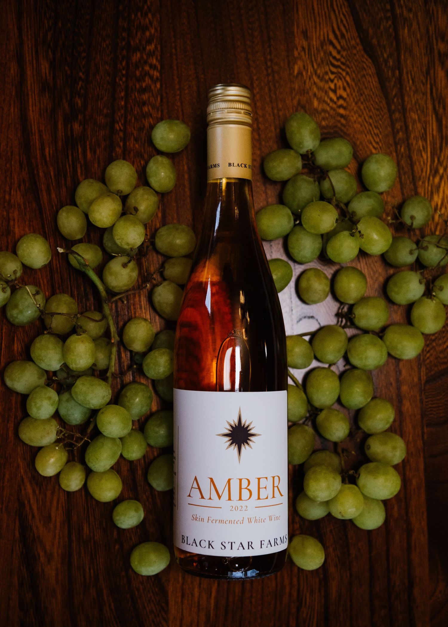 A bottle of Amber wine from Black Star Farms surrounded by fresh green grapes on a dark wooden surface.