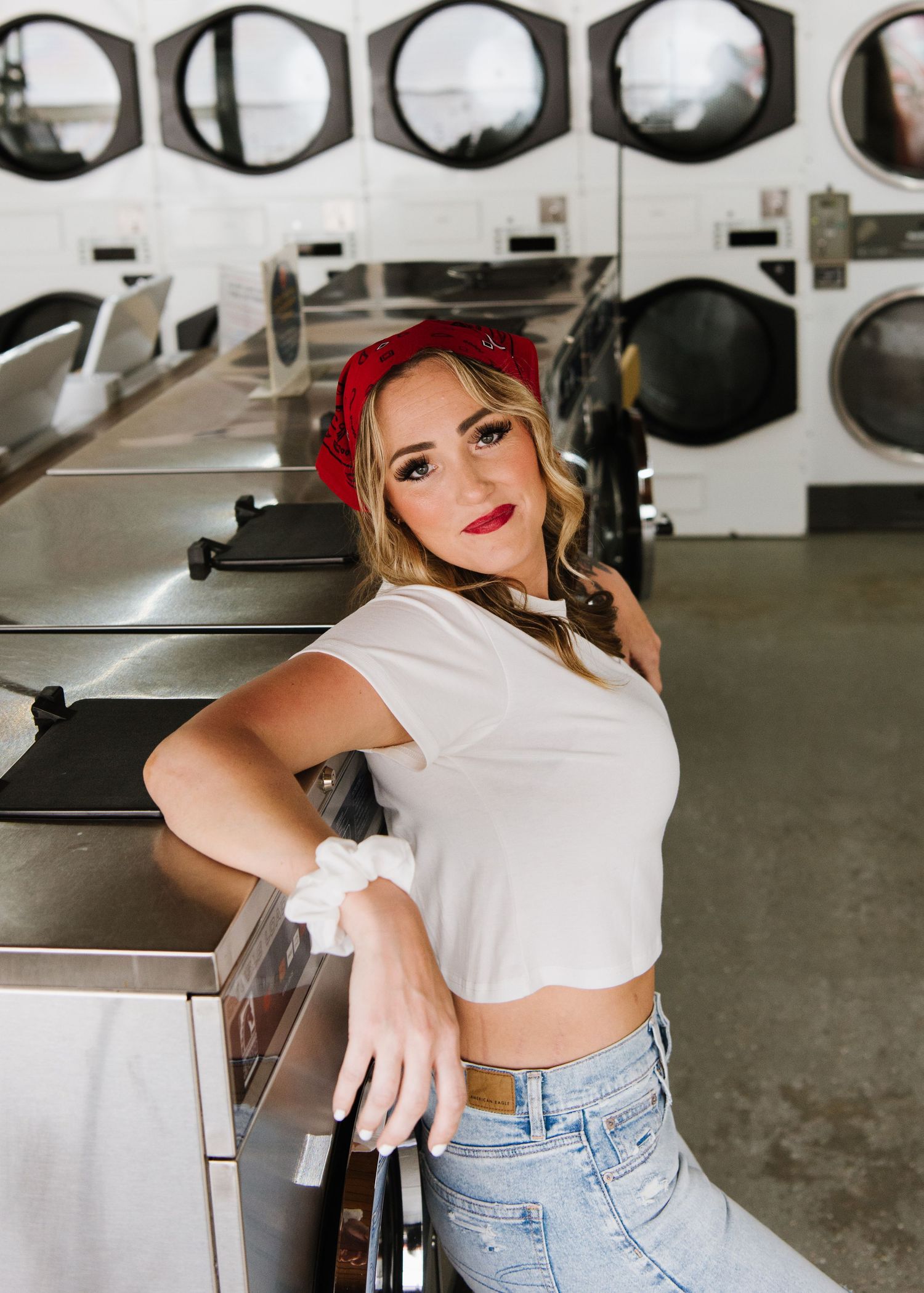 A person in a red bandana and white crop top poses inside a laundromat with washing machines in the background.