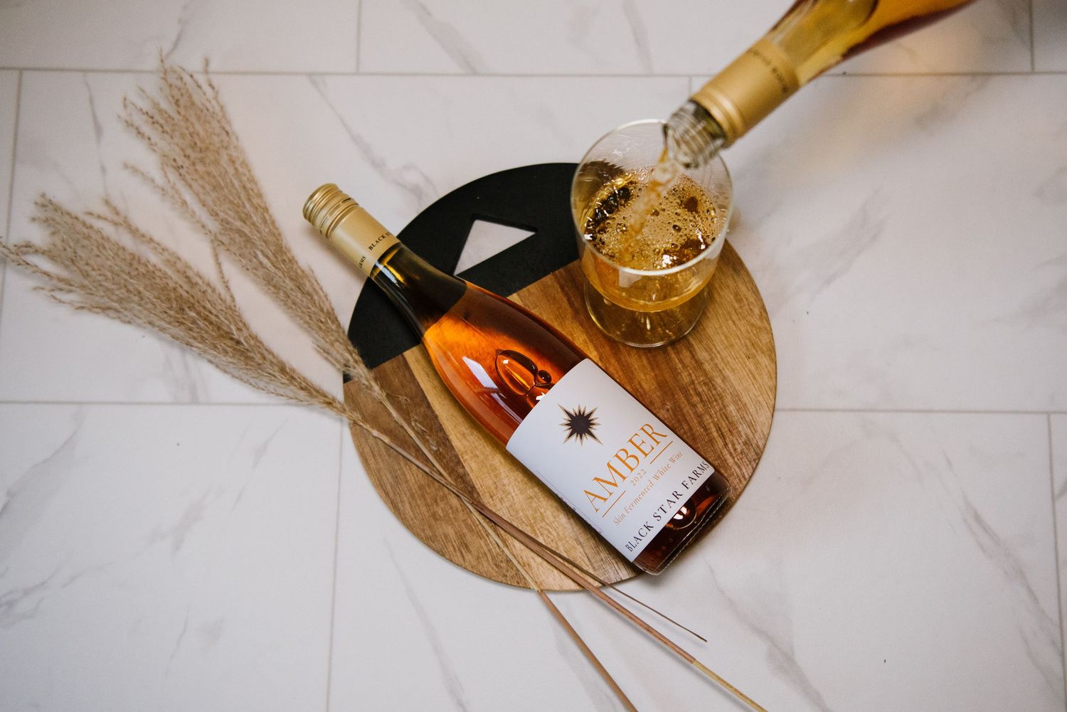 A wine bottle and glass on a wooden serving board with dried wheat stalks on white marble surface.