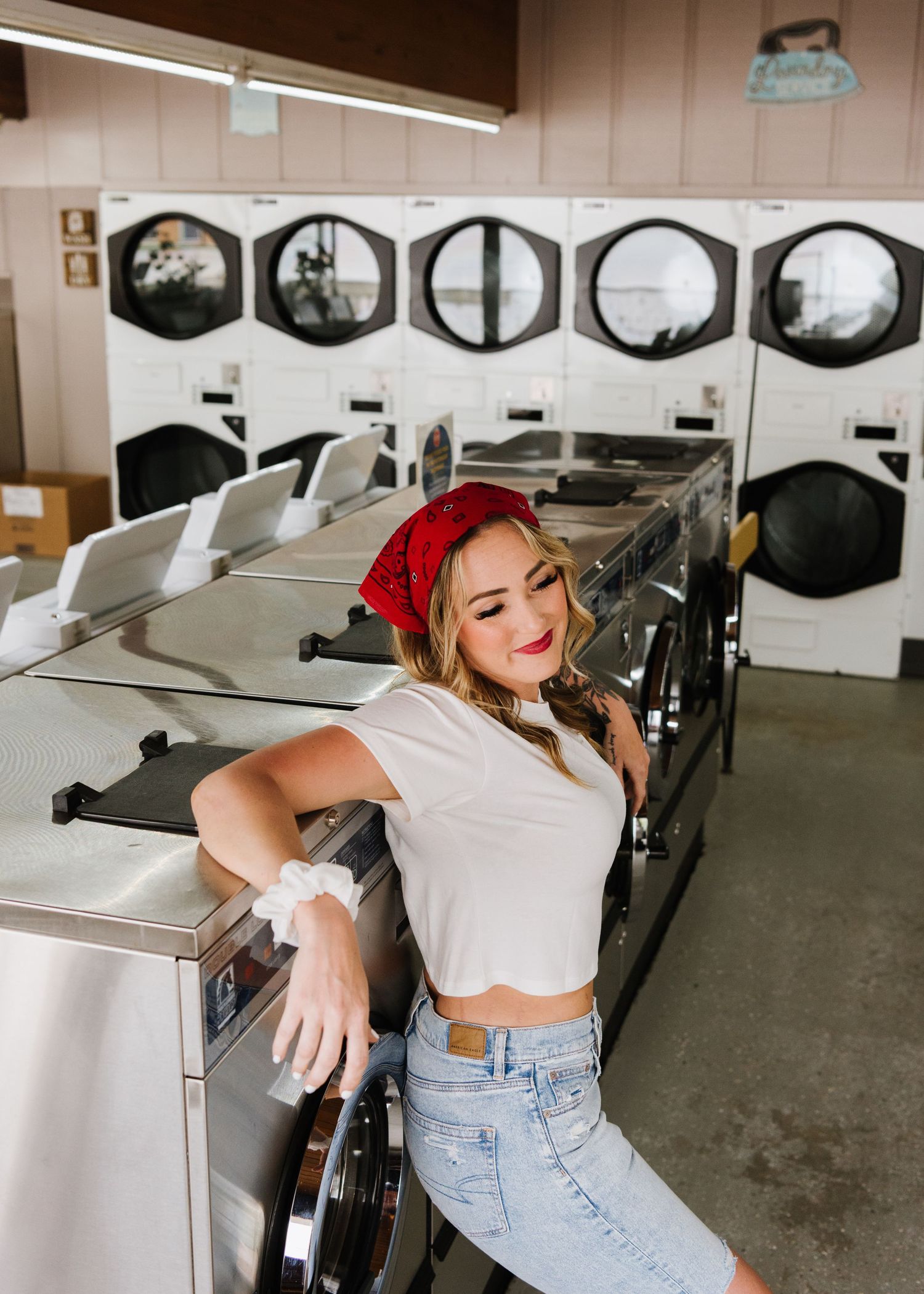 Fashion shoot with vintage laundromat setting shows someone in red bandana, white crop top and jeans posing by washing machines.