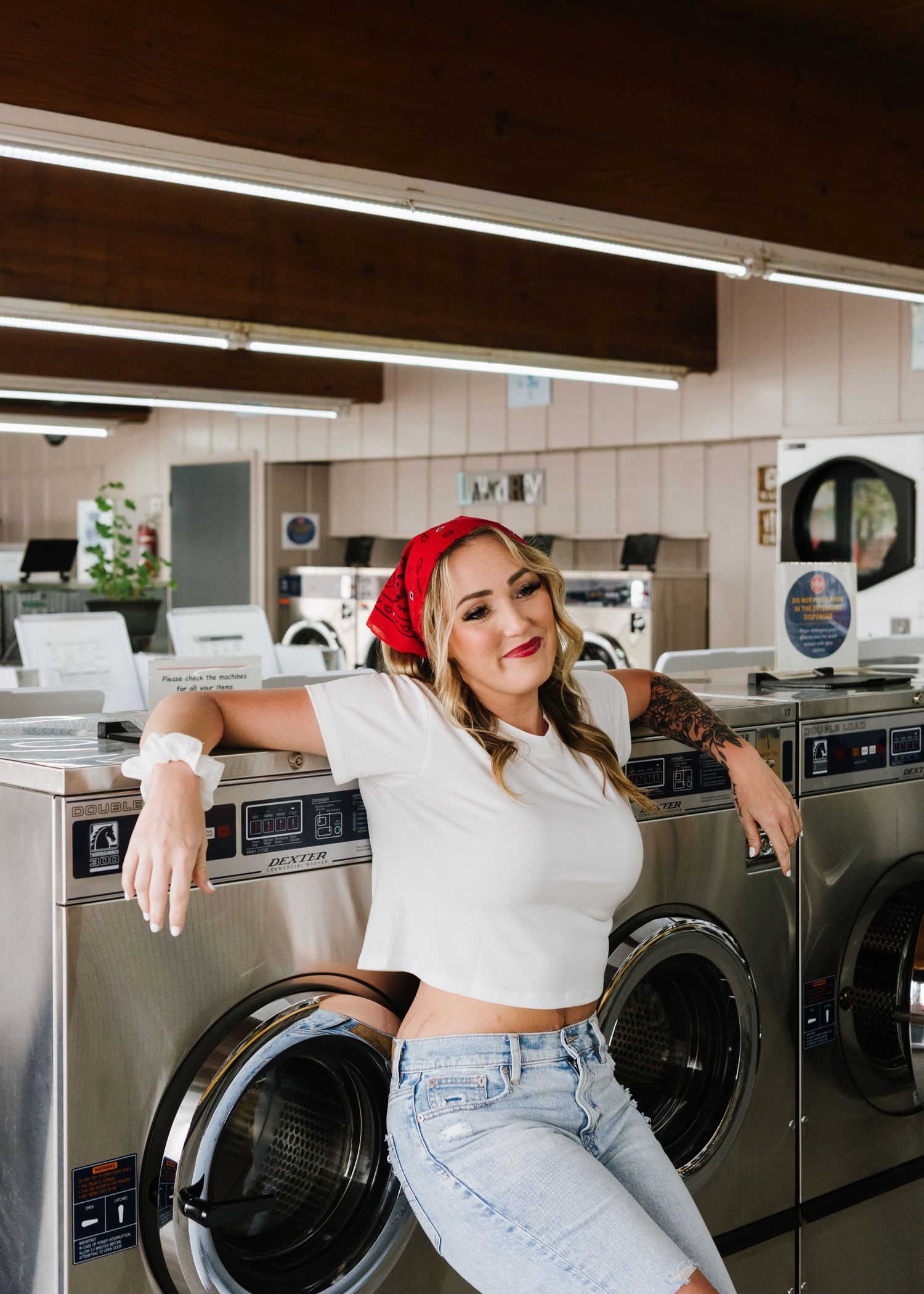 A woman wearing a red beanie and white crop top poses casually while leaning against a washing machine in a laundromat.