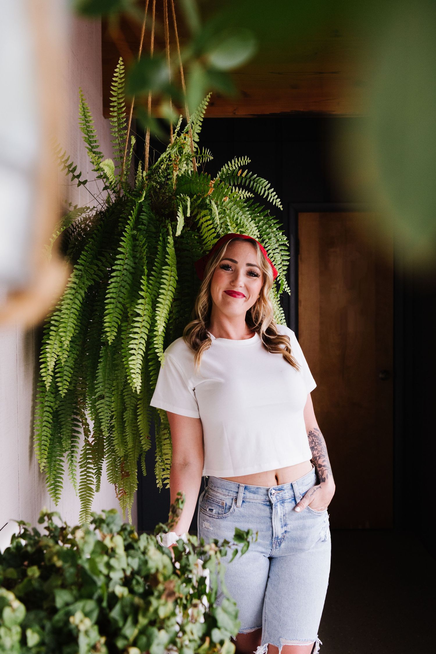 Woman in white crop top and denim shorts stands beneath a hanging fern plant in a naturally lit indoor setting.