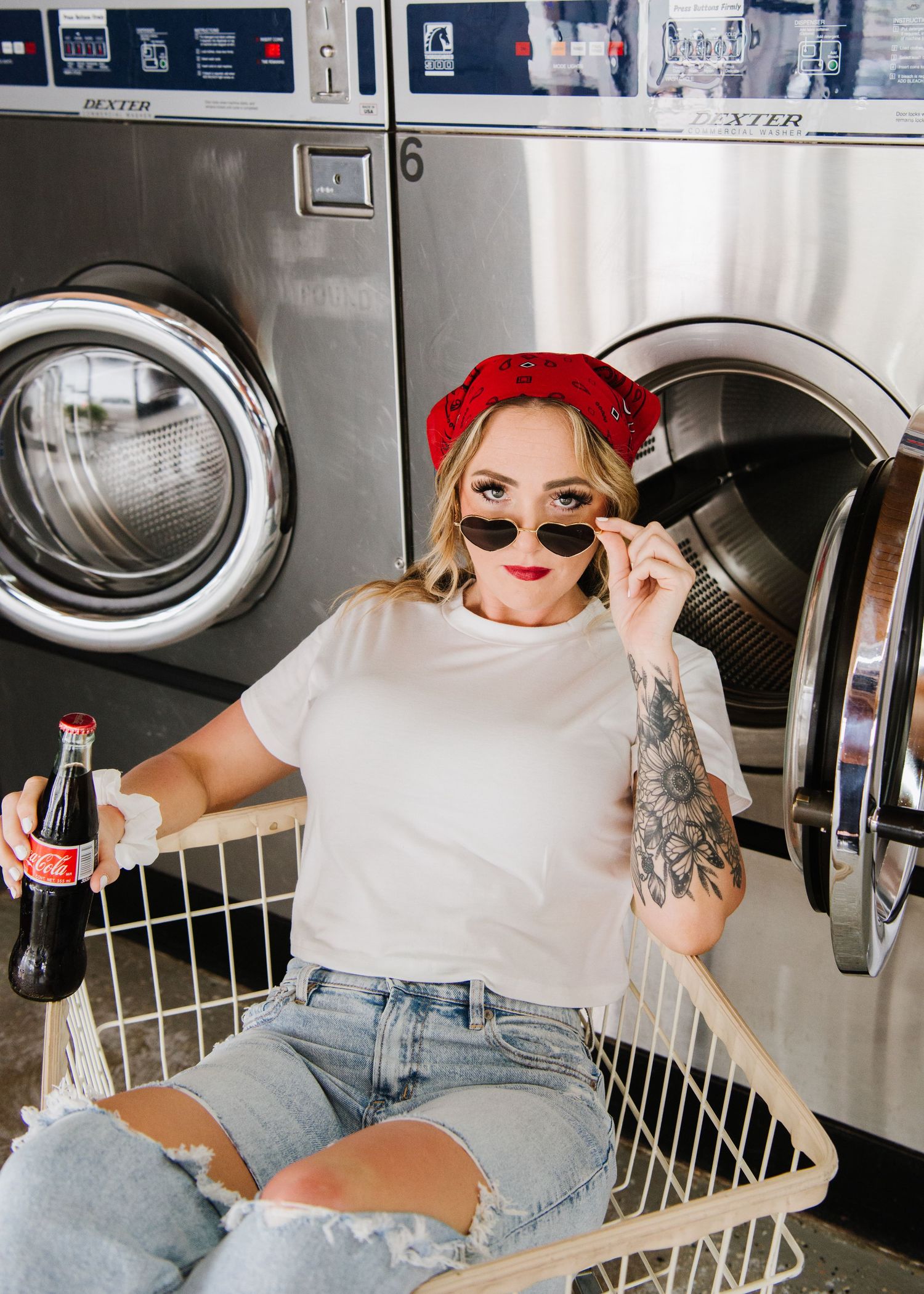 Young woman in sunglasses and red bandana relaxing at laundromat with a soda drink next to washing machines.