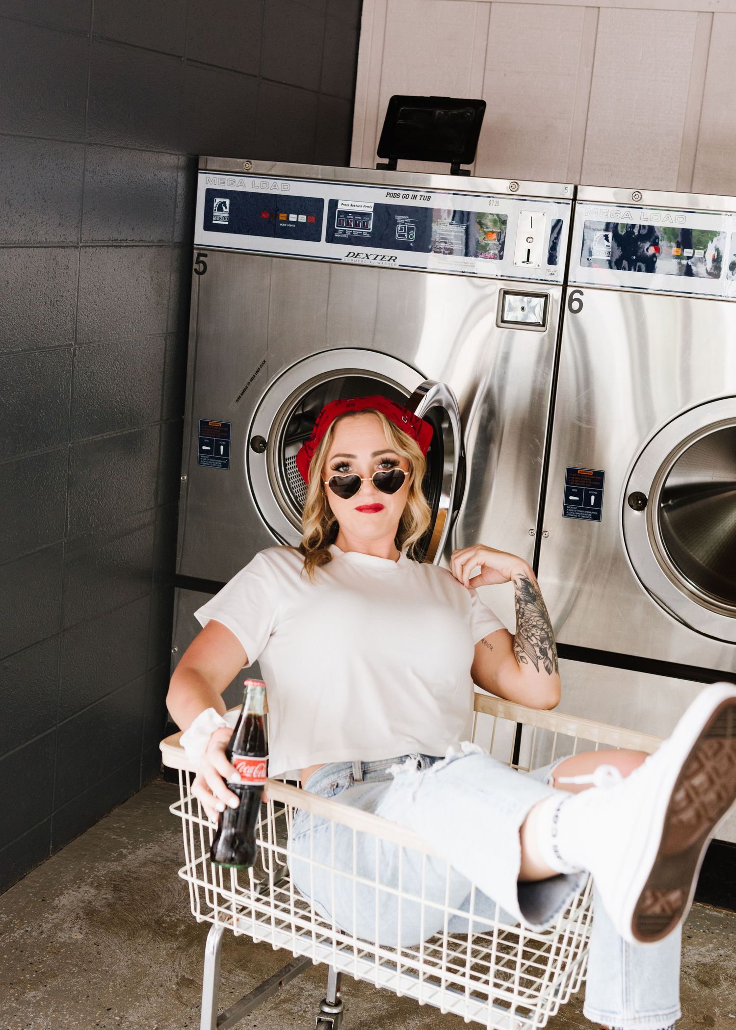 Trendy individual sits in laundry basket at laundromat wearing sunglasses and white t-shirt near industrial washers.