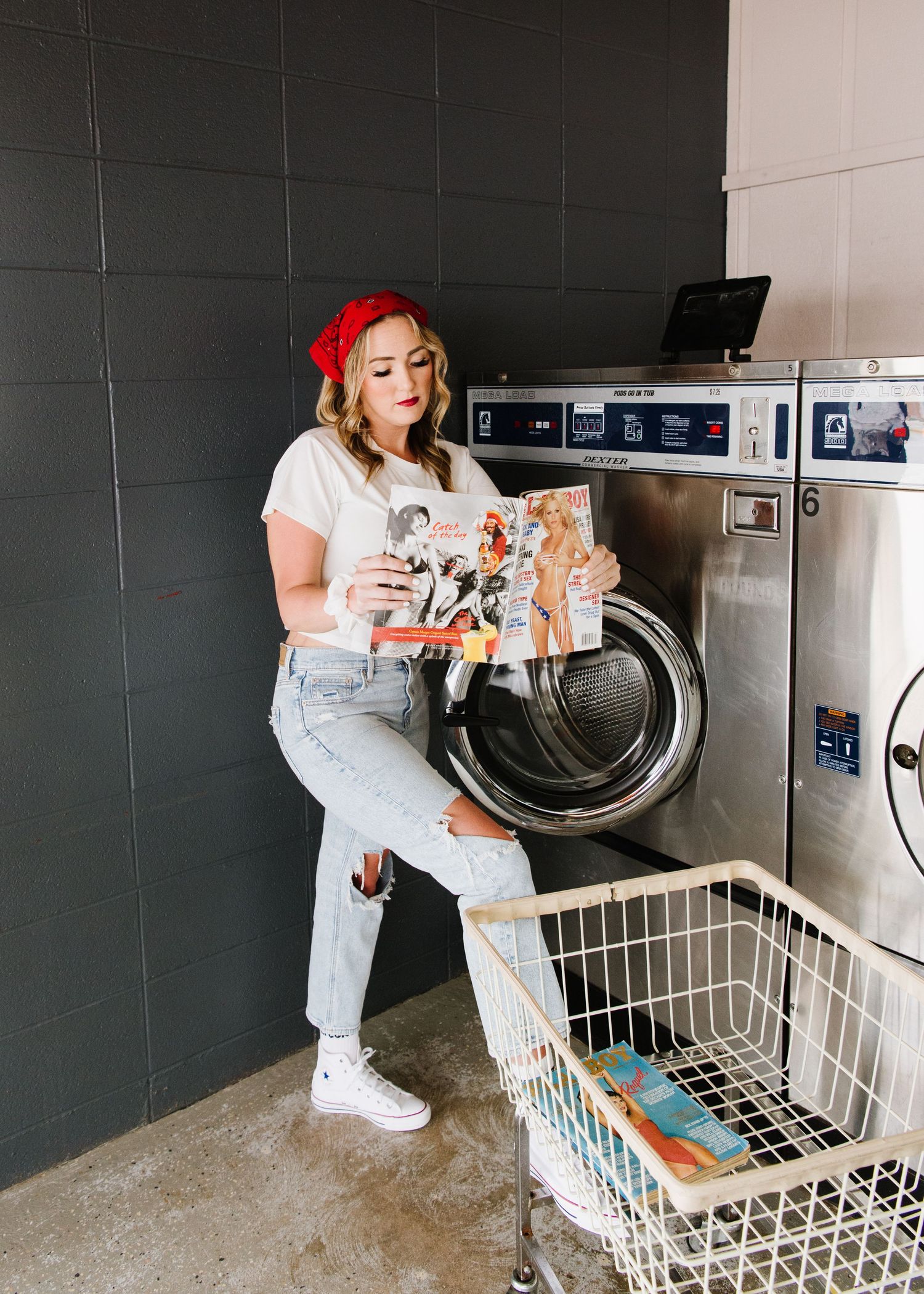 Someone in a red beanie and light jeans sits on a commercial washer while reading a manga comic at the laundromat.