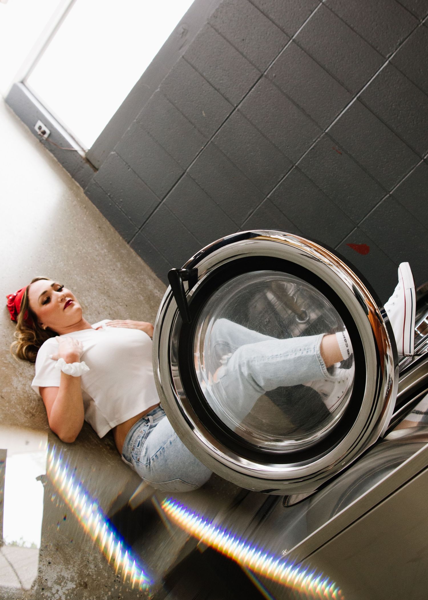 Person lying on floor next to front-loading washing machine in laundry room with modern black tile wall.