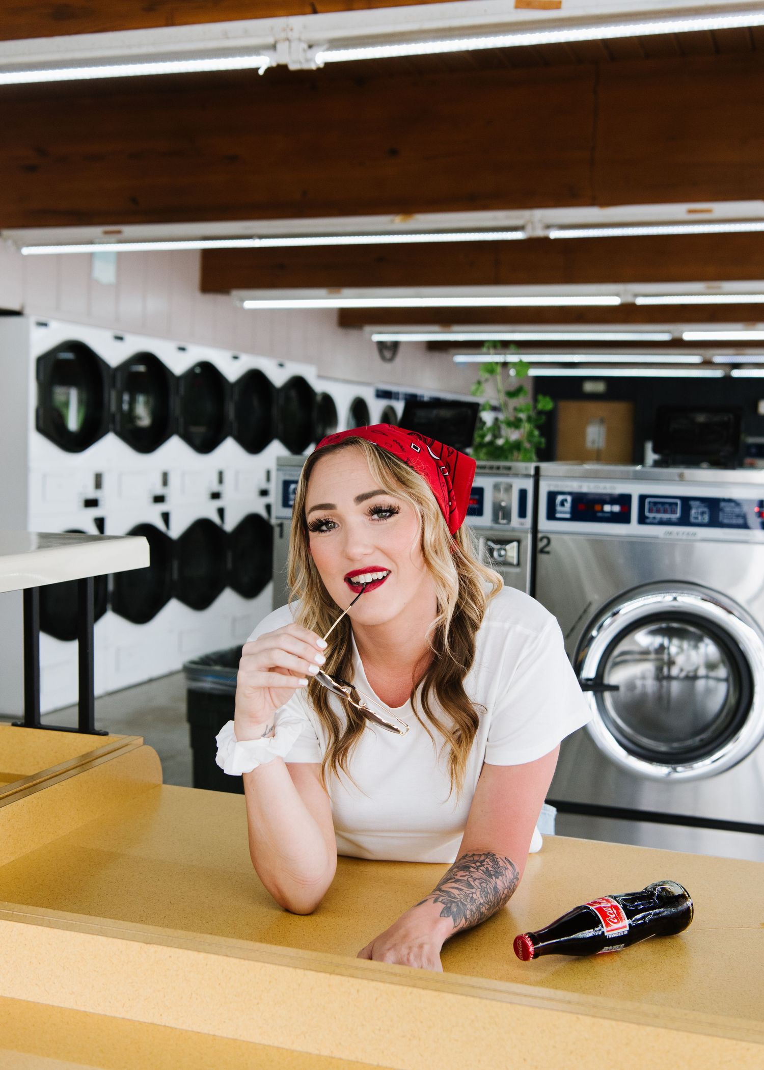 Woman in red bandana and white shirt leans on counter at retro-style laundromat with washing machines in background.
