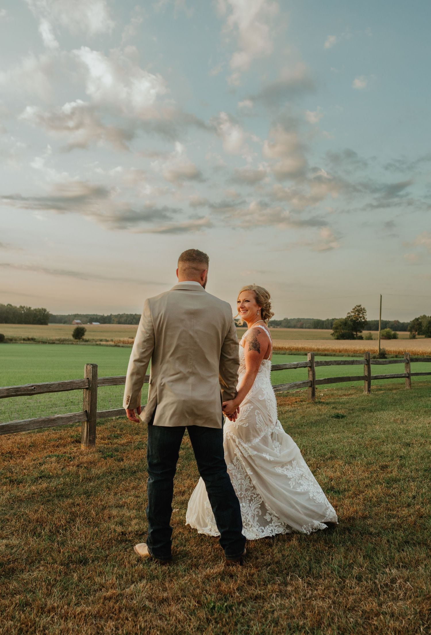 A couple in wedding attire stands on a grassy field at sunset with a rustic wooden fence in the background.