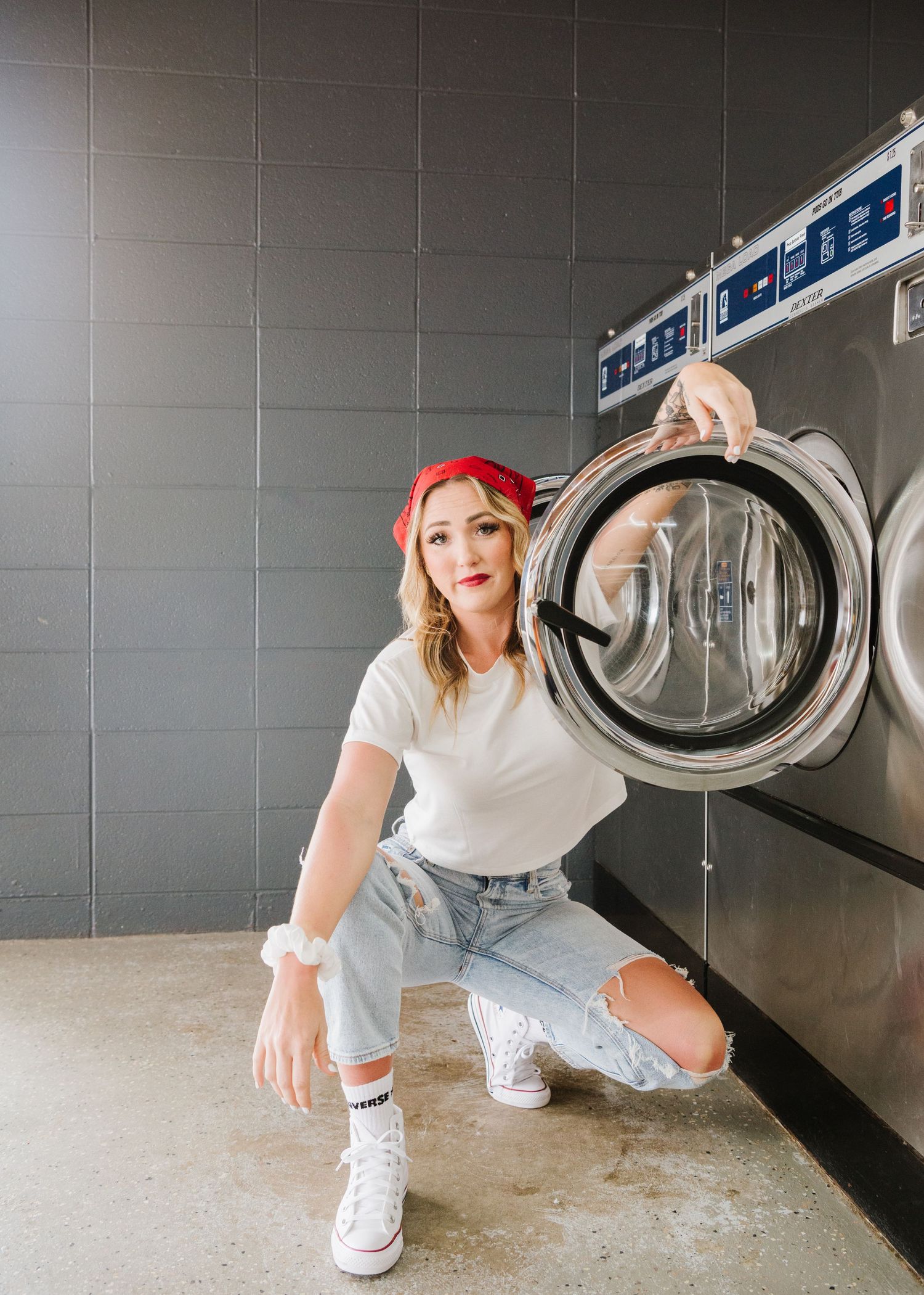 Person in white t-shirt and ripped jeans poses in front of commercial washing machines at laundromat.