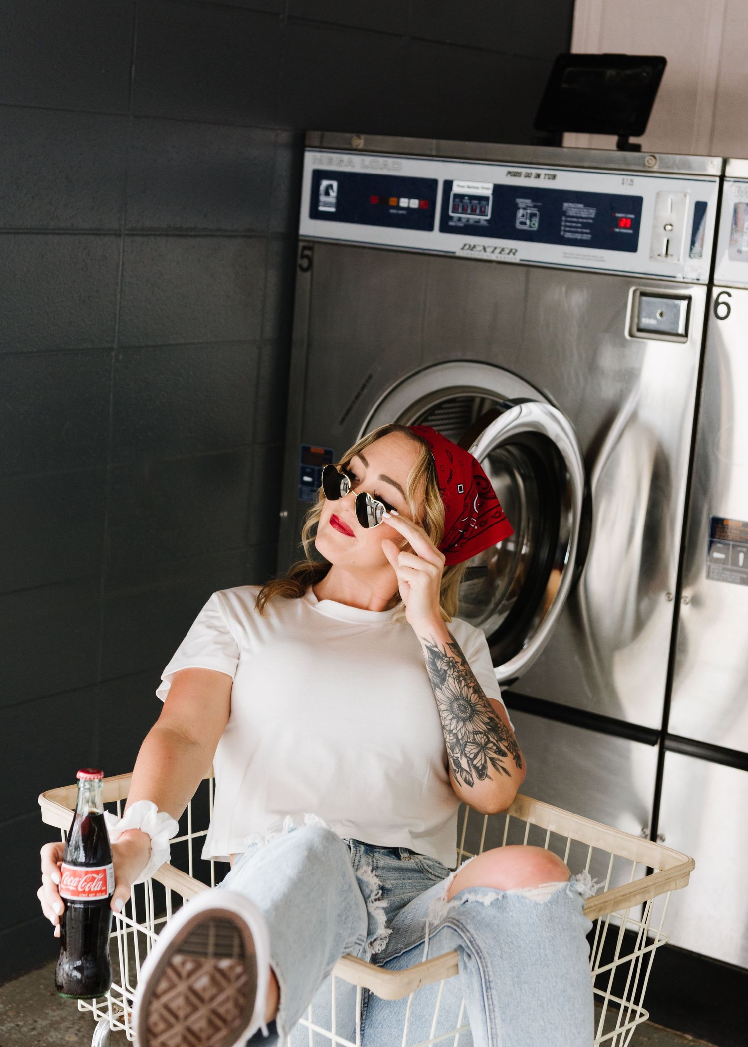 A tattooed person in a white shirt sits atop a laundry basket at a laundromat while wearing red headphones.
