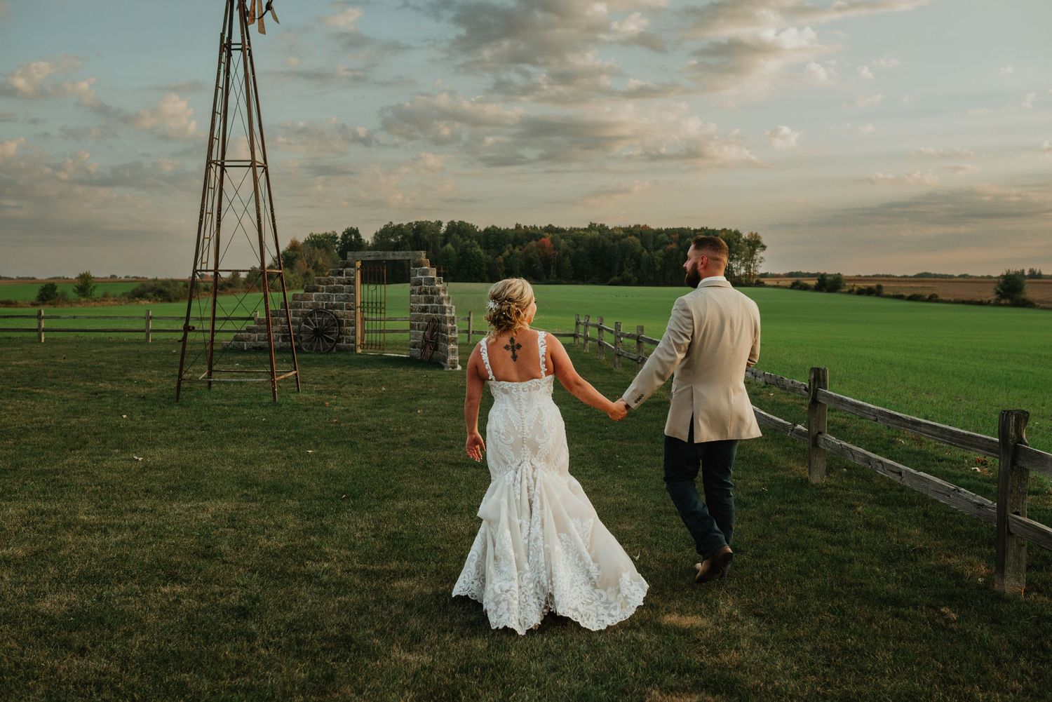A couple holds hands in a rustic farm setting at sunset with a windmill and wooden fence in the background.