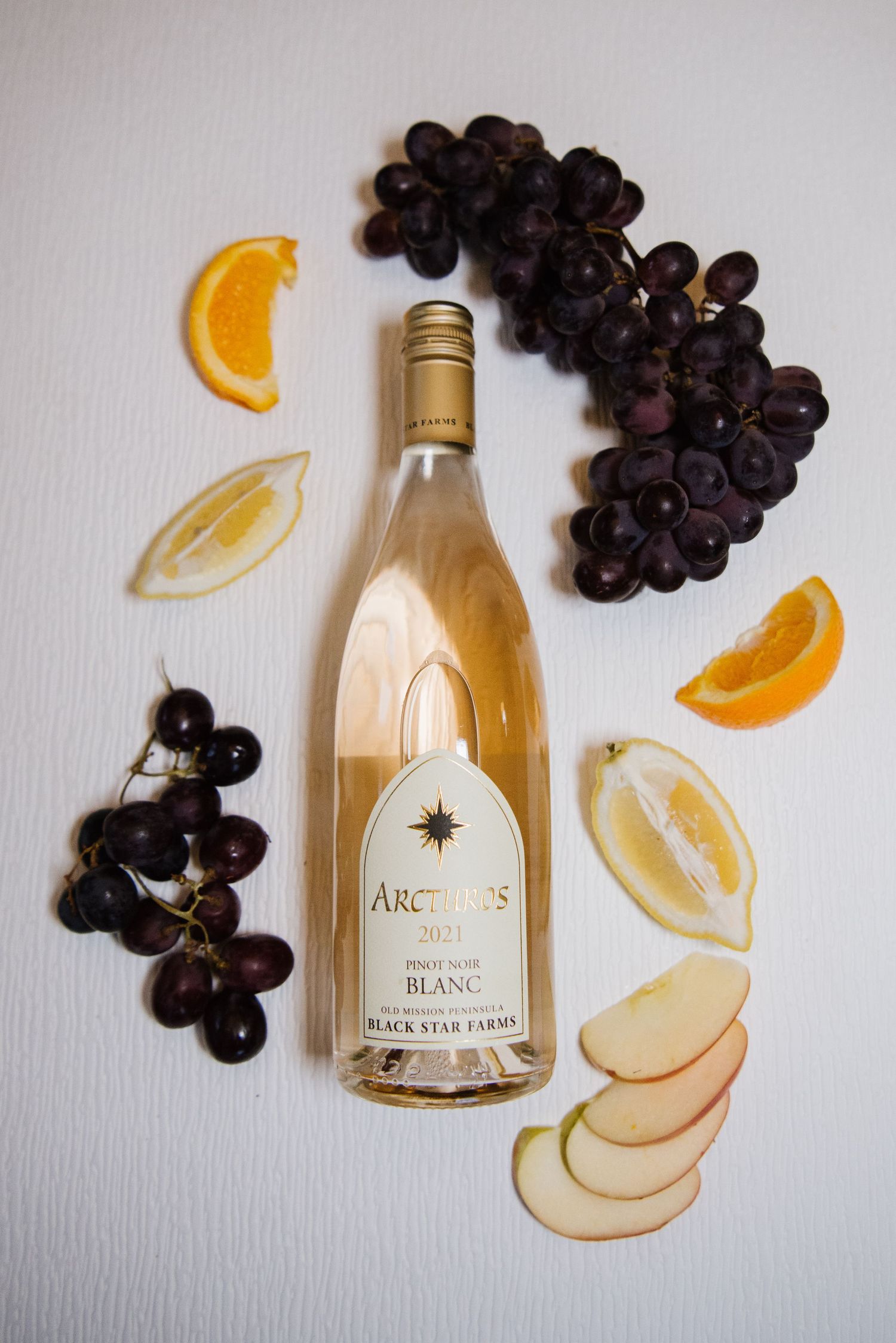 A bottle of white wine surrounded by fresh grapes, orange slices, and apple wedges on a white surface.