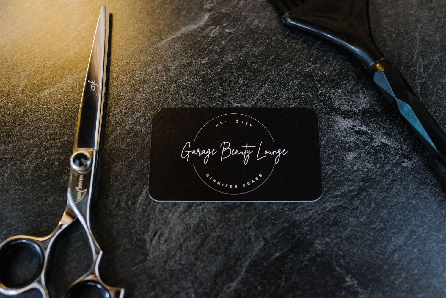 Professional hairdressing scissors and black business card on dark stone surface with dramatic lighting.
