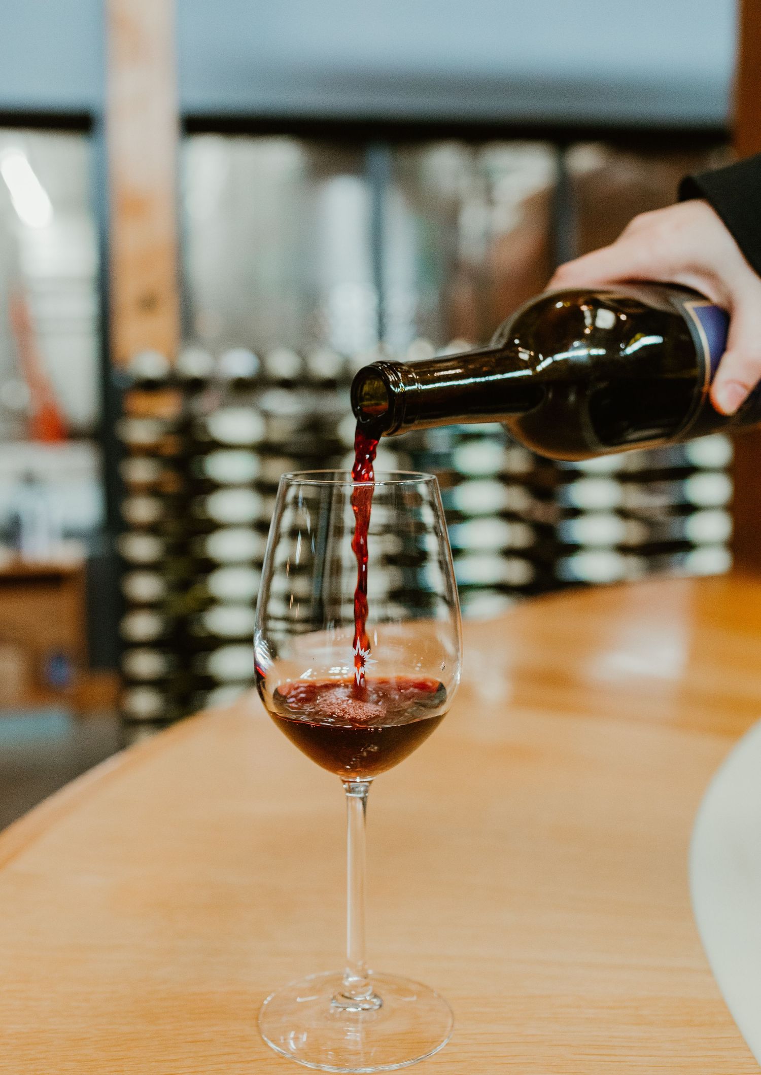 Red wine being poured from a bottle into a wine glass on a wooden bar counter with wine racks in background.