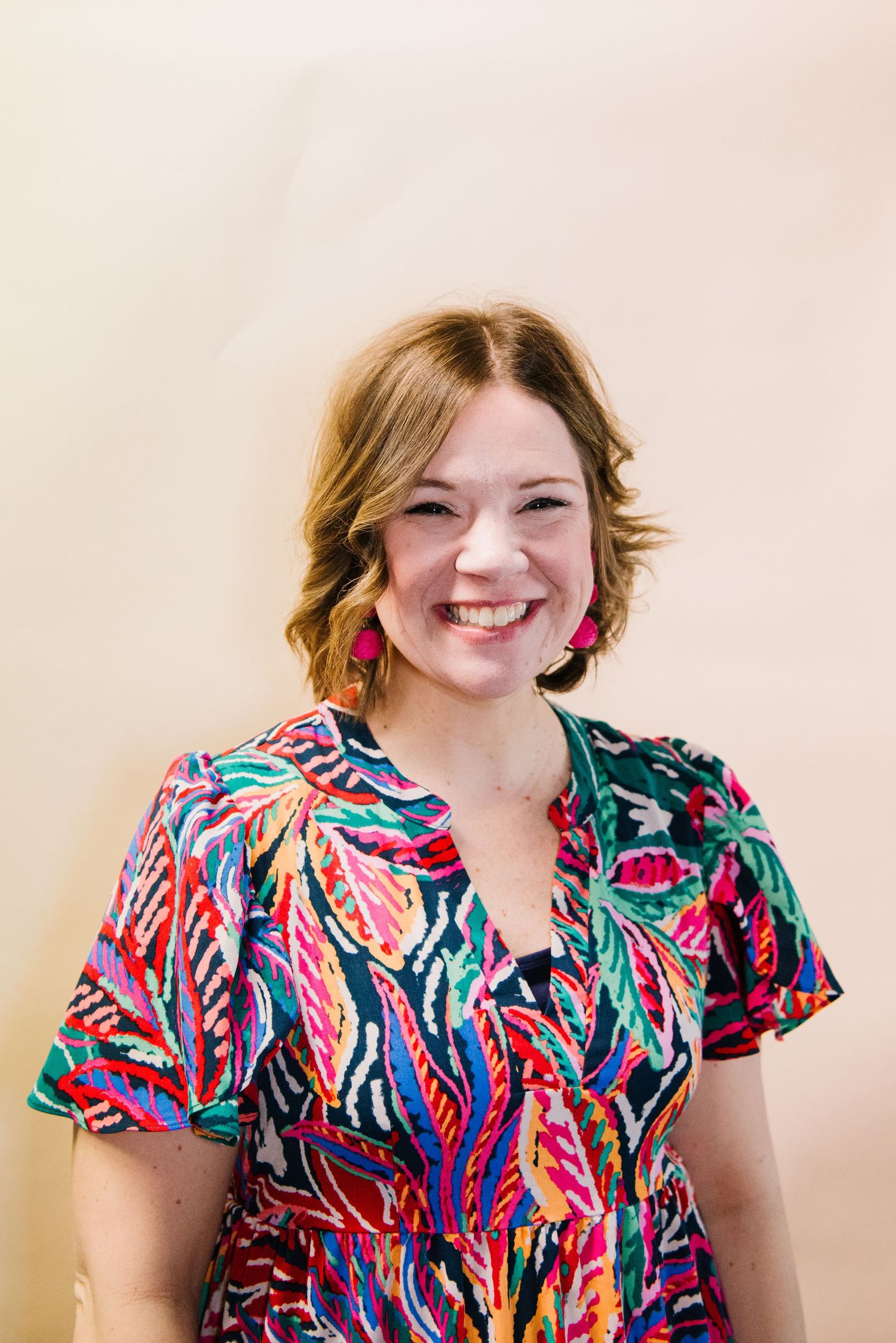 Portrait of someone in a vibrant tropical print dress with flutter sleeves and colorful dangling earrings against a light background.