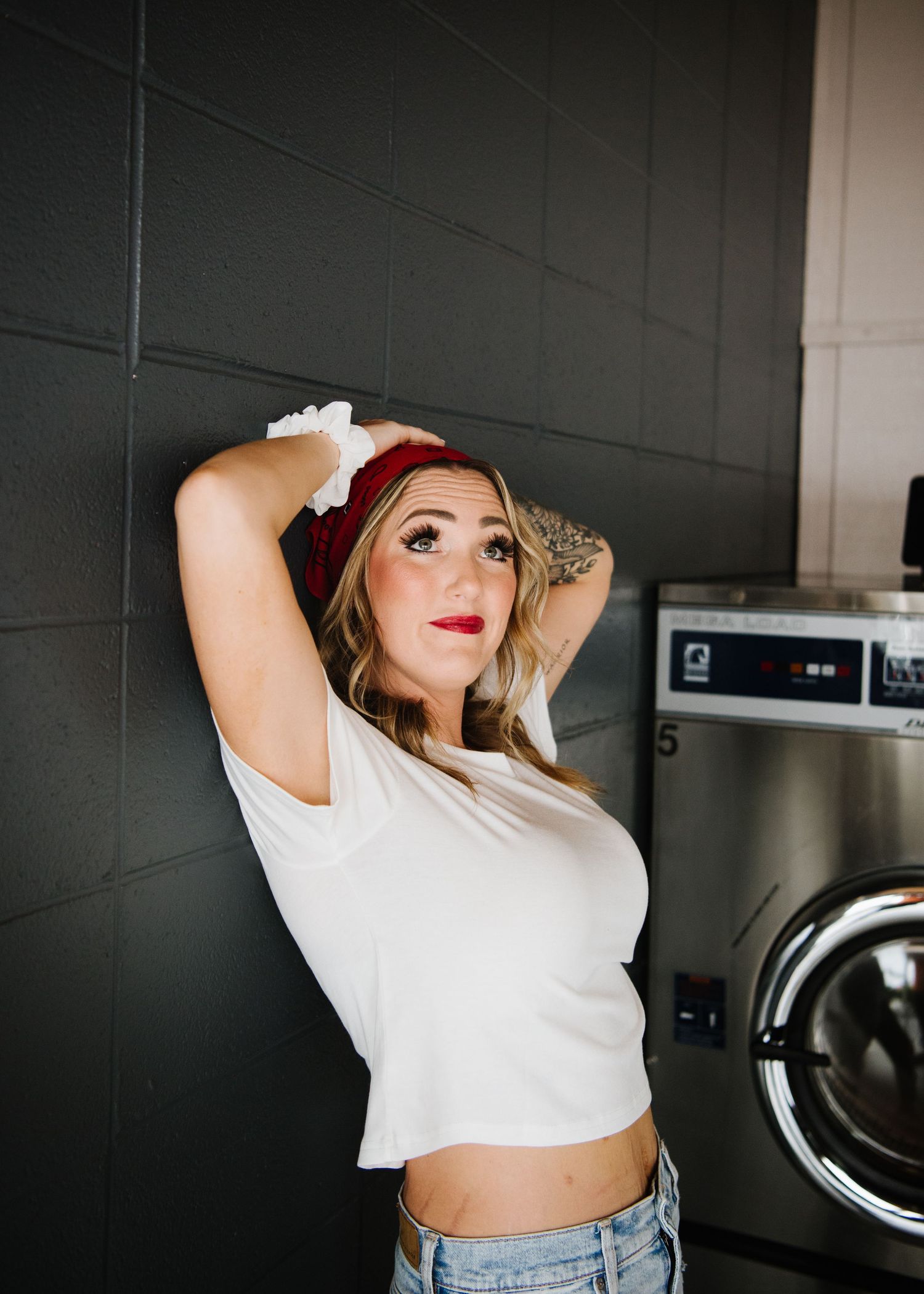 Fashionable woman in white crop top and red headband poses by washing machine in laundromat.