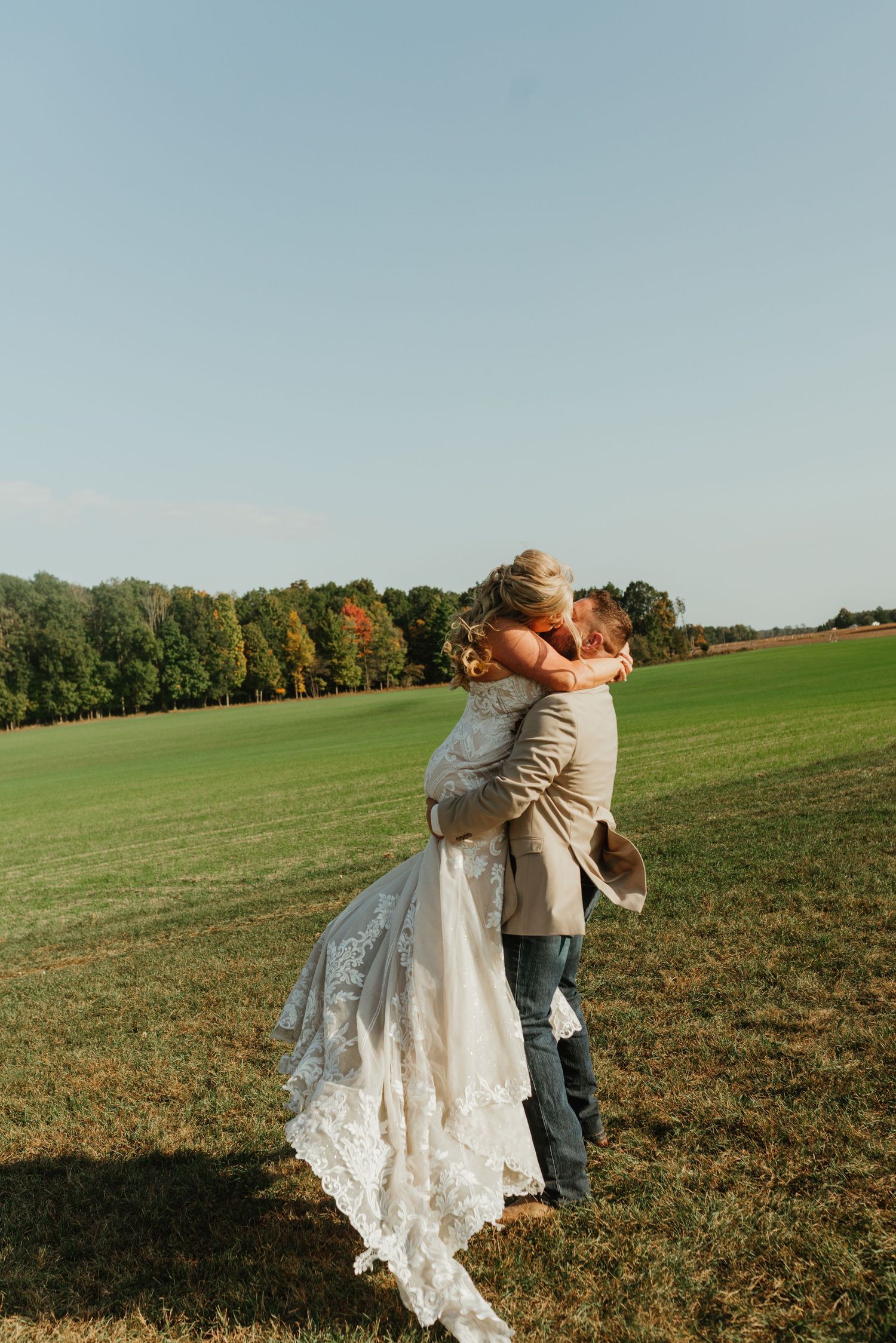 Couple embraces in an outdoor field with autumn trees and blue sky in the background.
