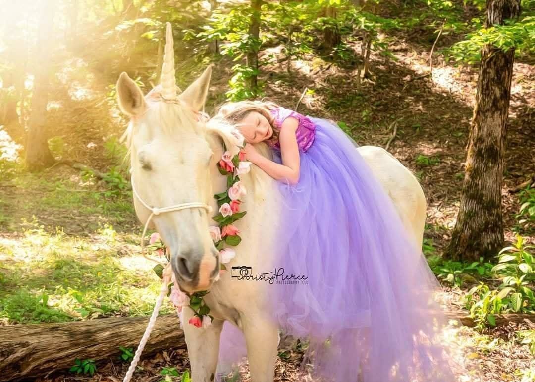 A dreamy fairy tale scene in a sunlit forest featuring a white unicorn and flowing purple tulle gown.