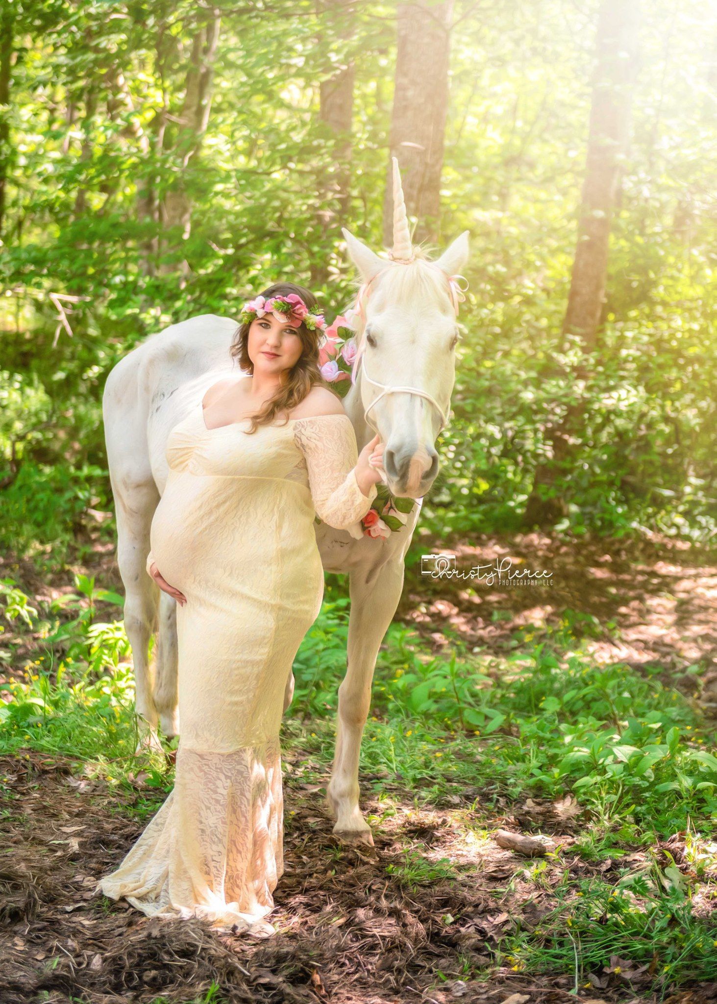 Ethereal maternity portrait in a sunlit forest featuring a cream lace dress and white horse companion.