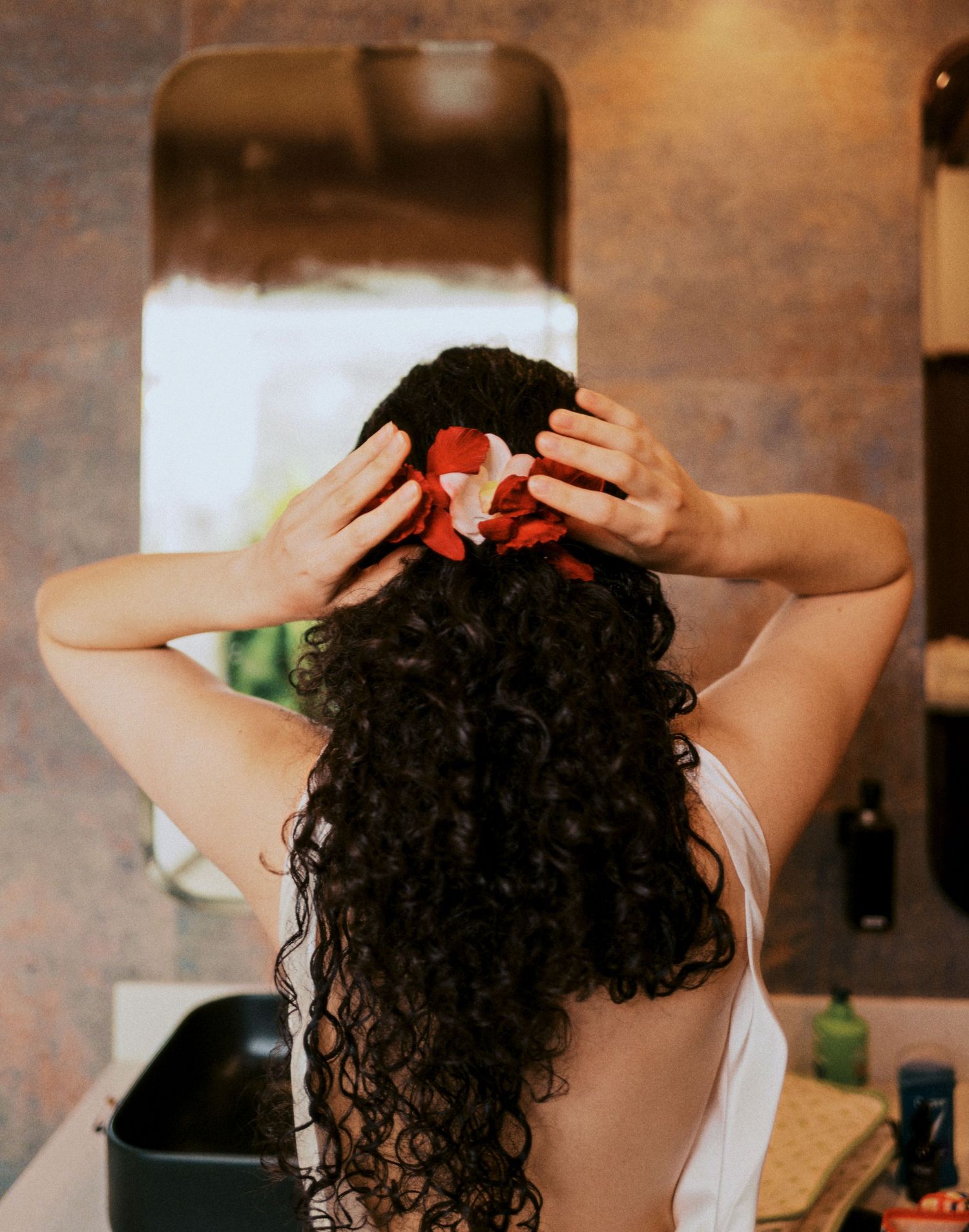 Person with long curly dark hair wearing a white top seen from behind while placing their hands on their head.