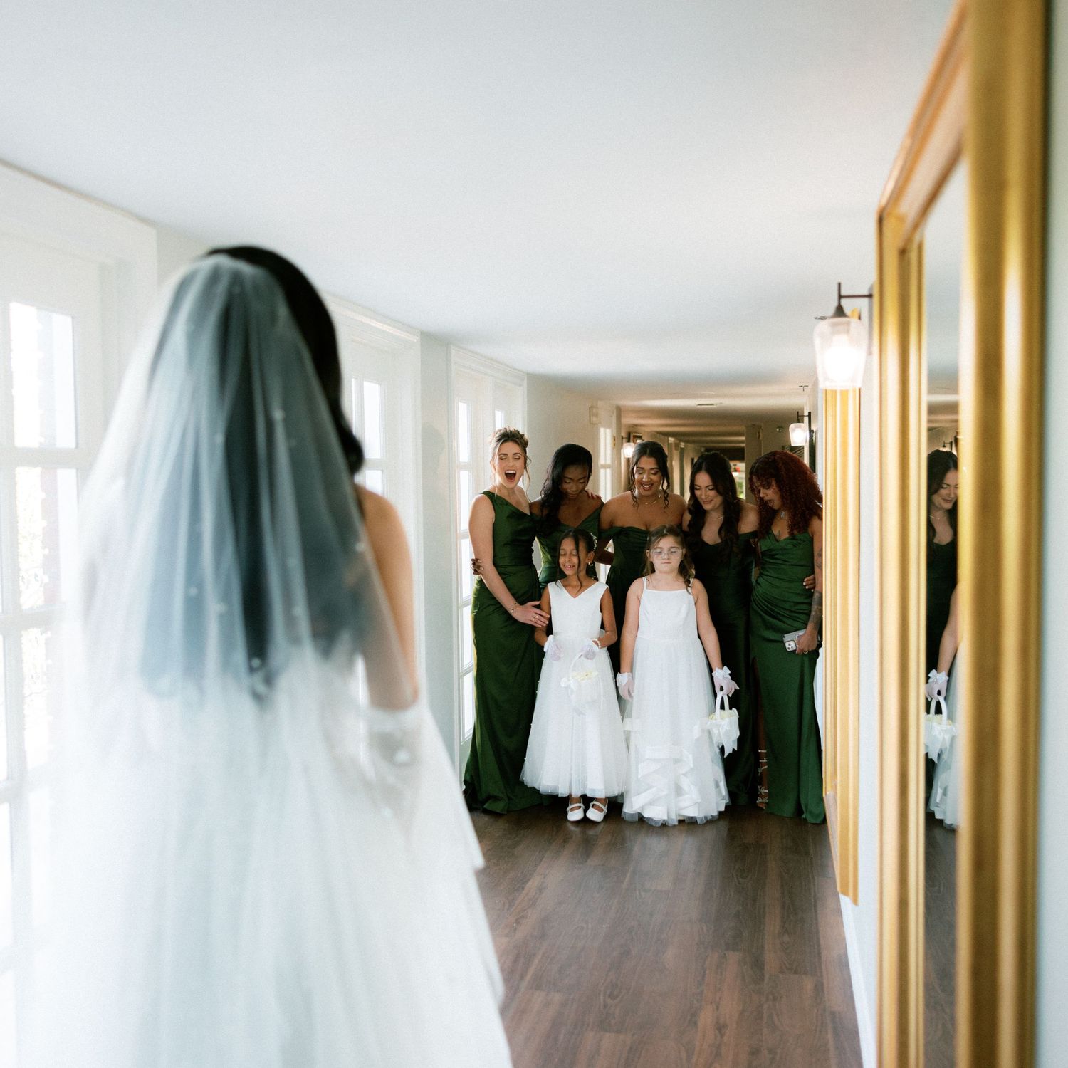 Wedding party gathering in a bright hallway with white walls and hardwood floors.