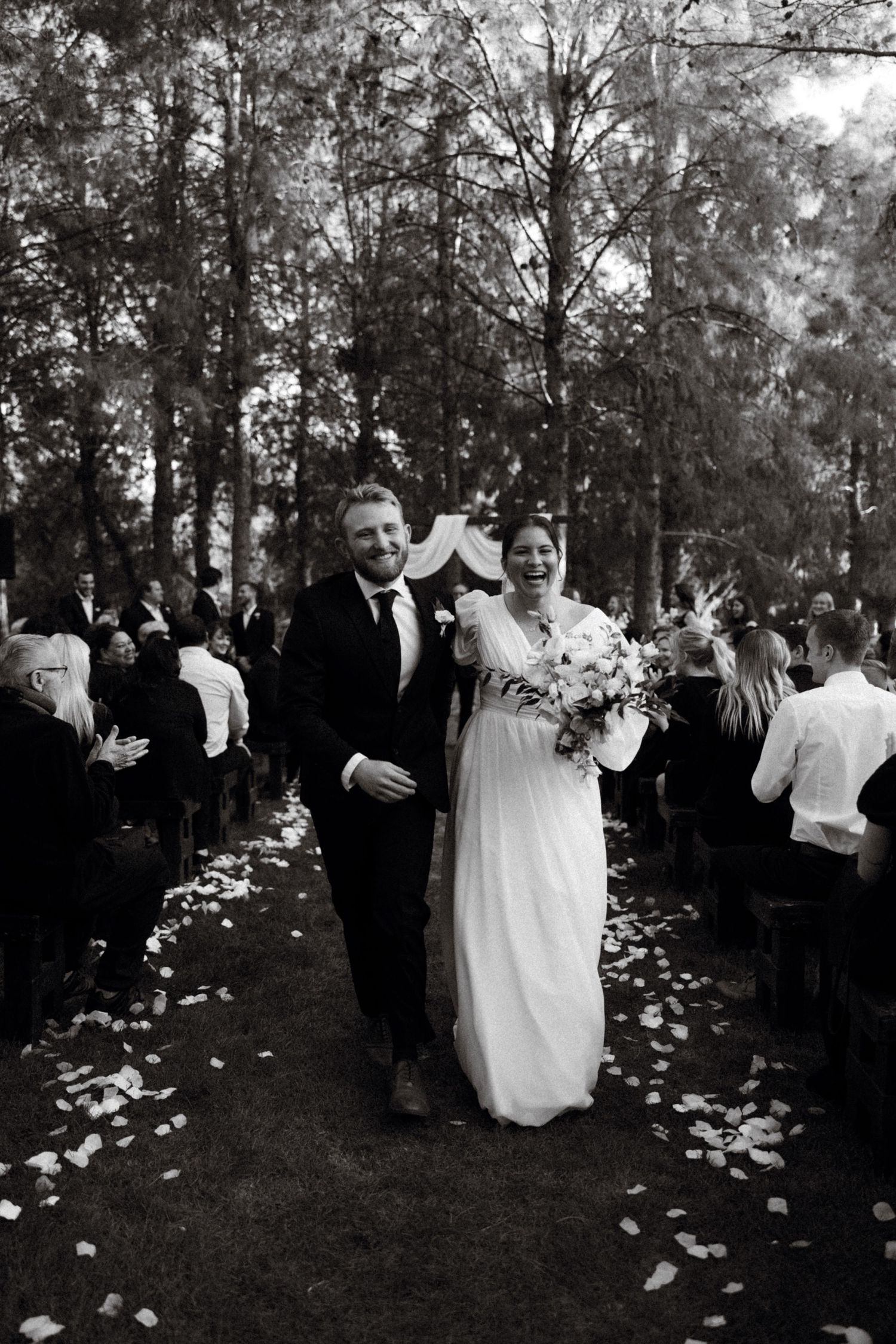 A couple walking down a flower-petal covered aisle through a wooded ceremony site.