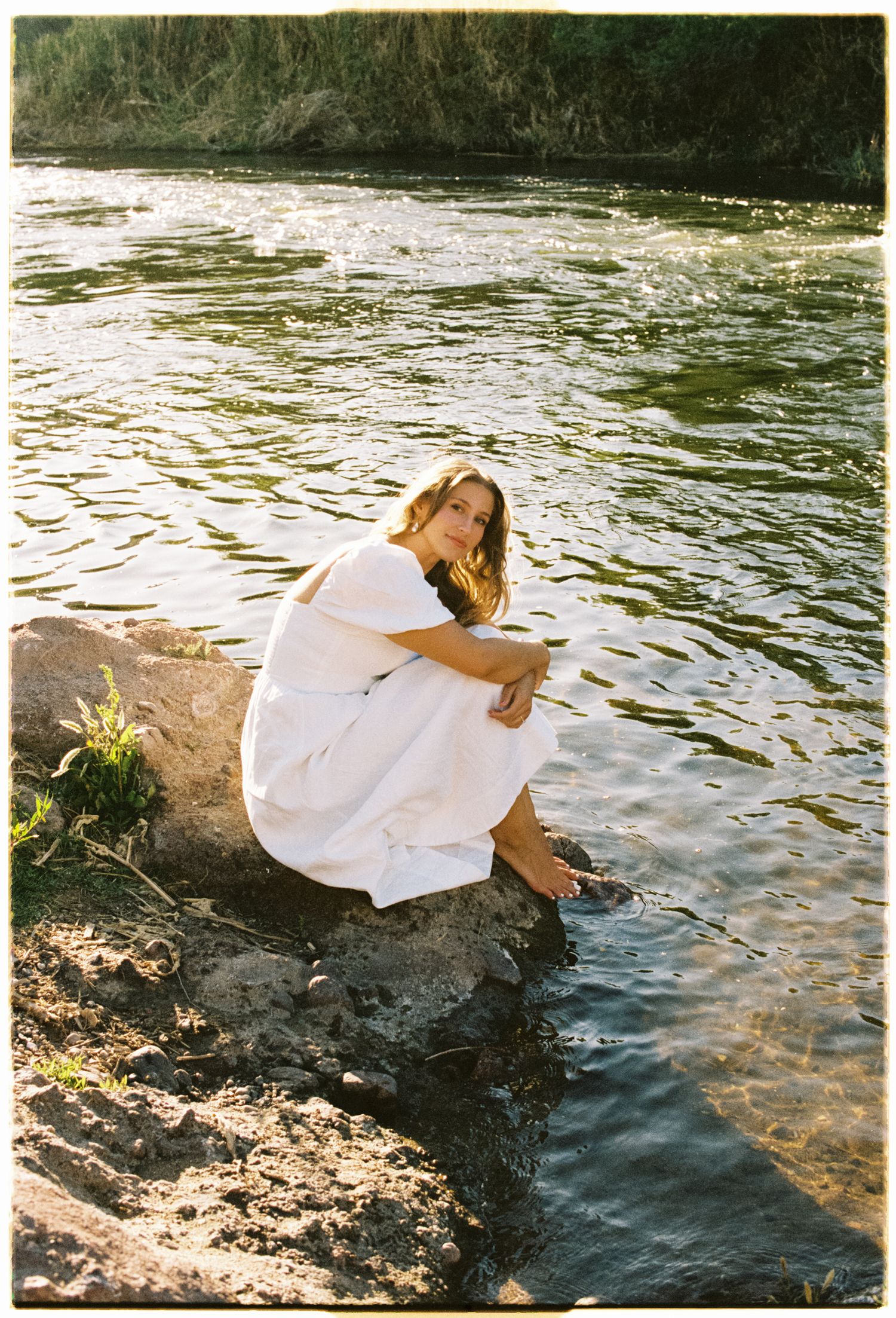 A person in a white dress sits on rocky riverside, gazing at peaceful flowing water on a sunny day.