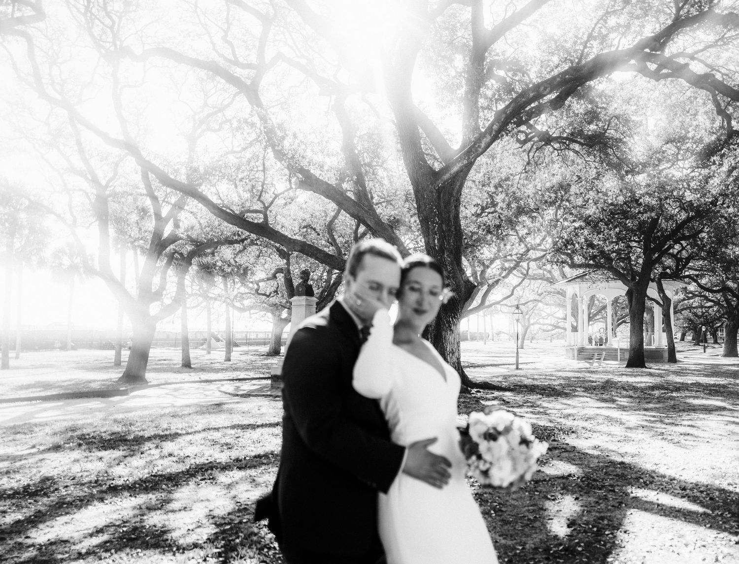 A bride and groom embrace under sunlit oak trees in a dreamy black and white wedding photograph at a park.
