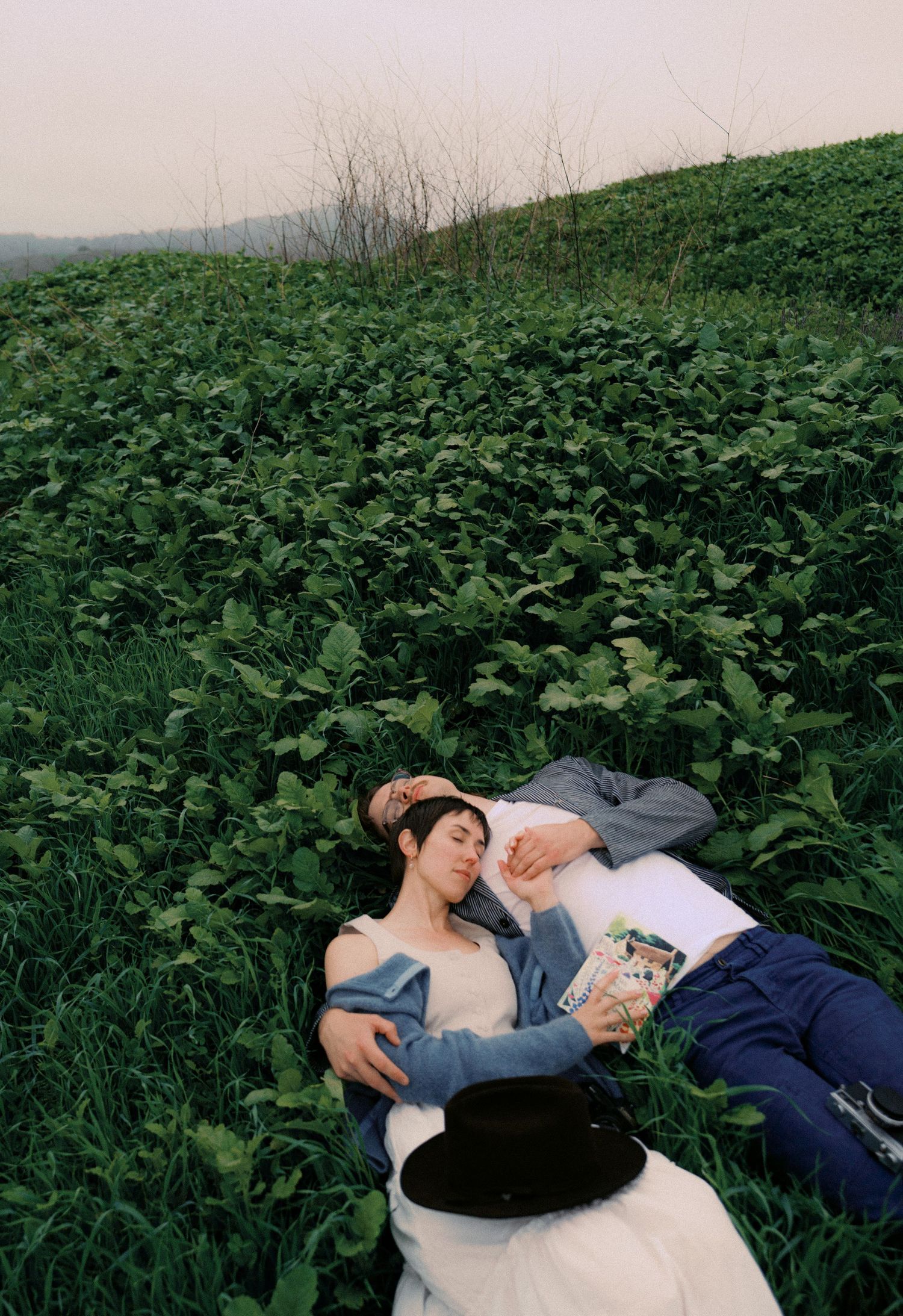 Two people lie peacefully among lush green foliage on a hillside during a hazy afternoon.