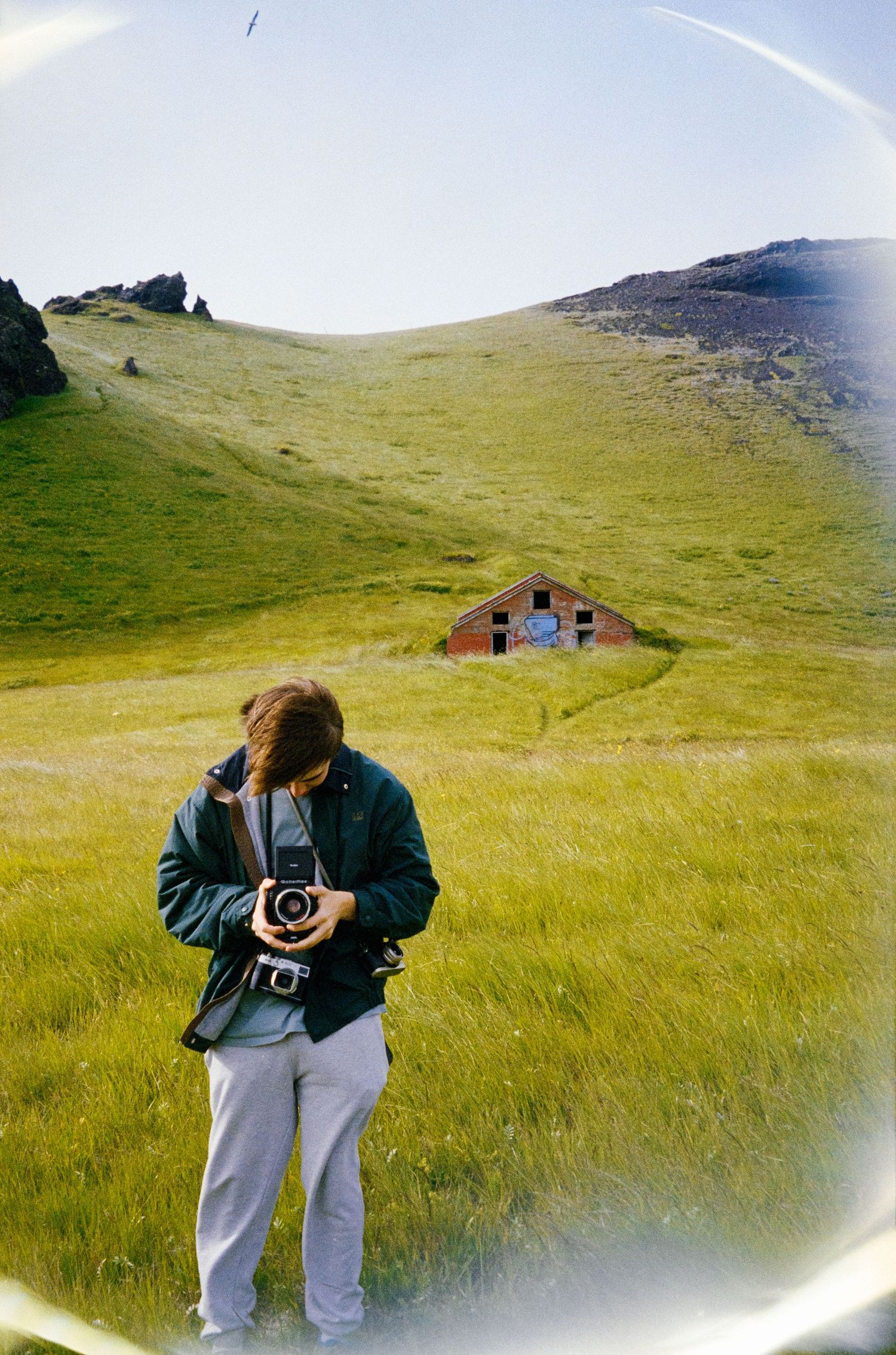 A photographer with vintage camera on grassy hillside with small buildings in distance.