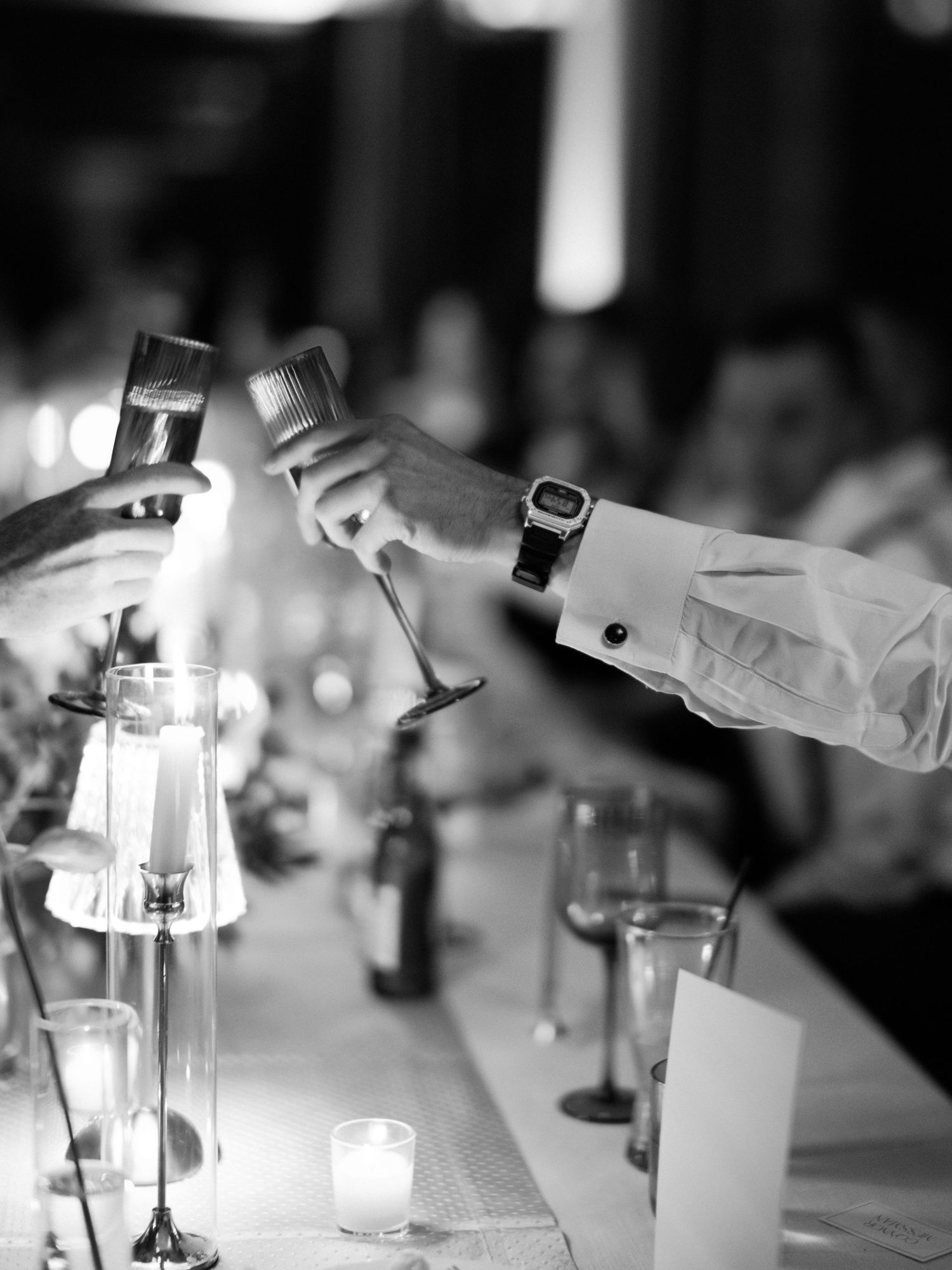 Black and white photo of hands toasting with champagne glasses at an elegant dinner table with candlelight.