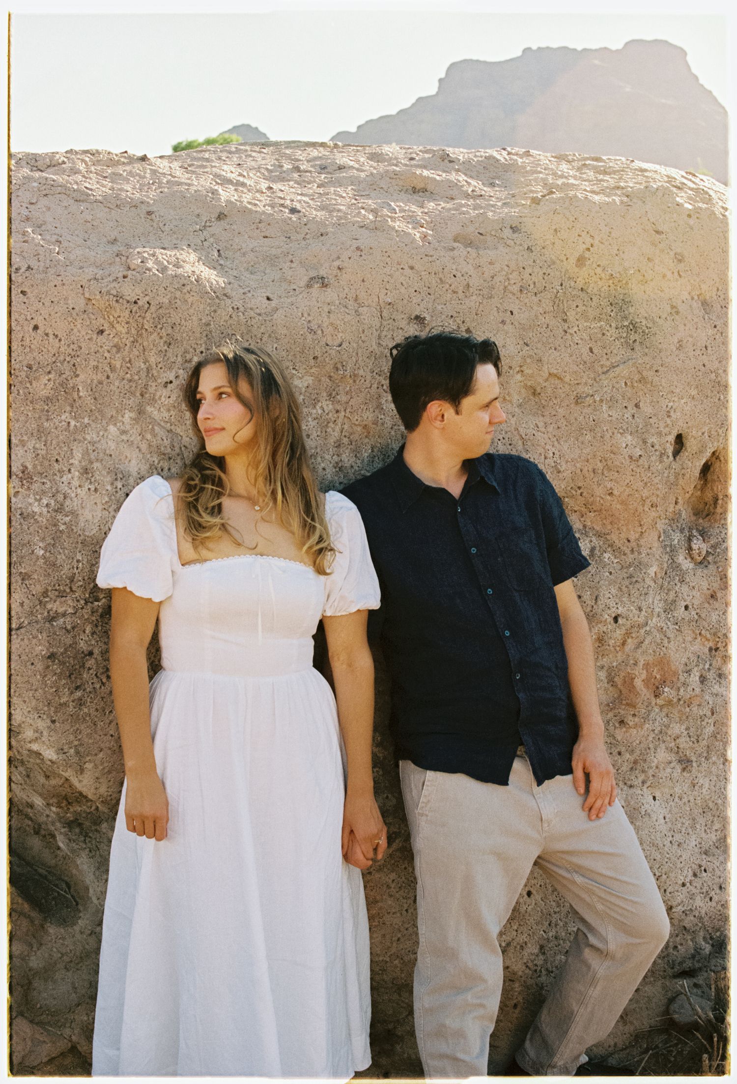 A couple poses against a rocky wall, with woman in white dress and man in navy shirt and khaki pants at golden hour.