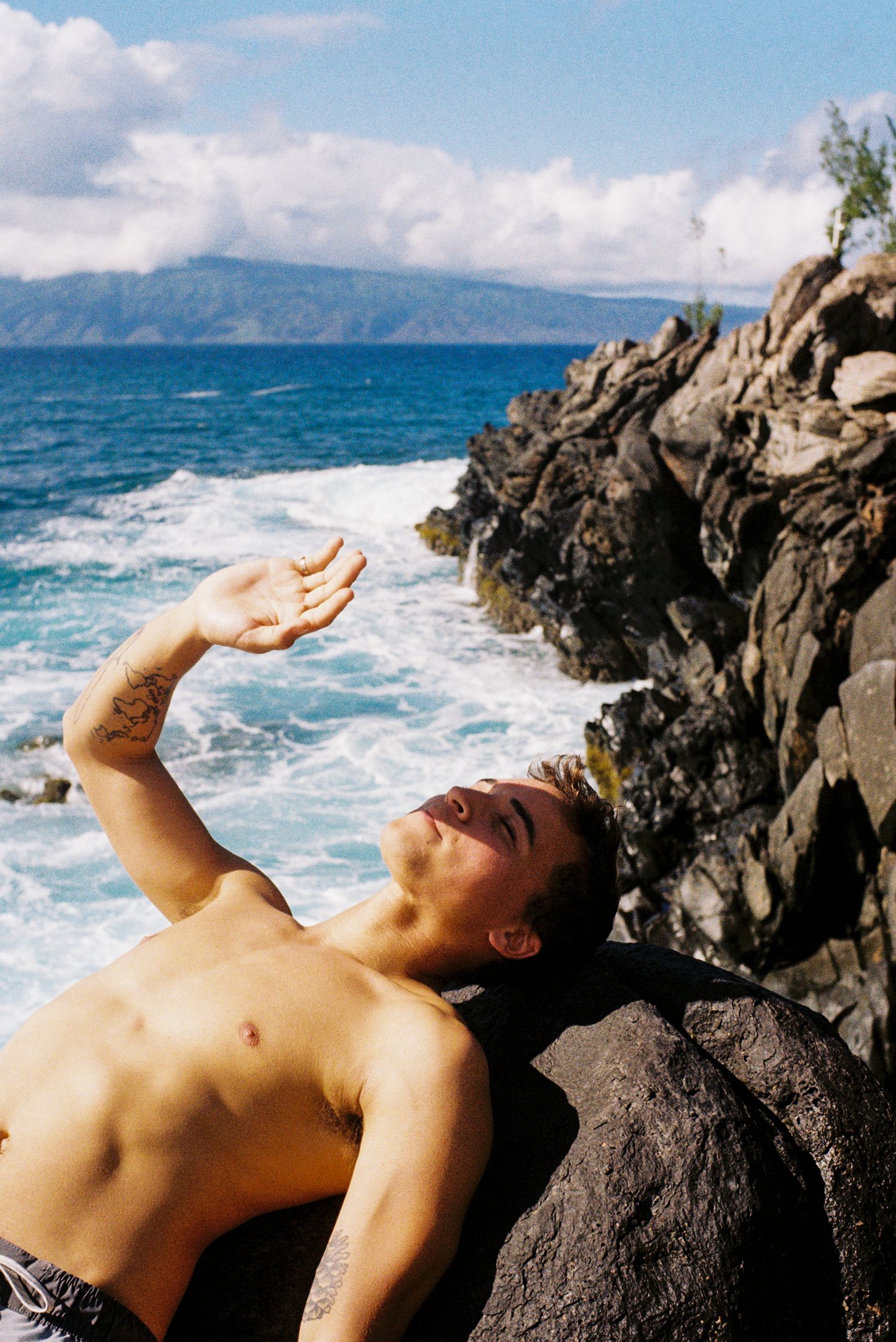 Shirtless person reclining on coastal rocks with dramatic ocean waves crashing behind.