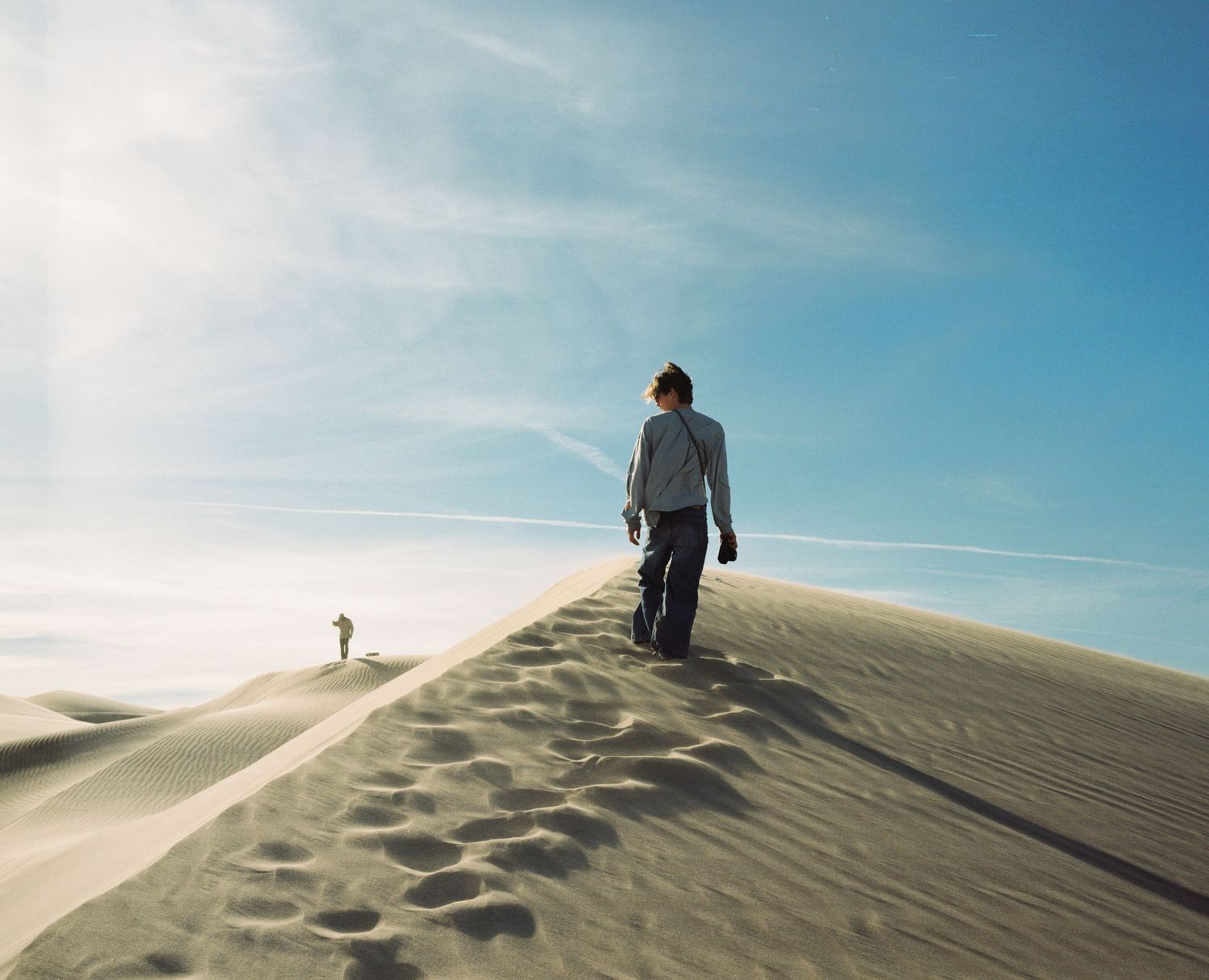 Silhouetted figure walking along sand dune ridge against bright blue sky.