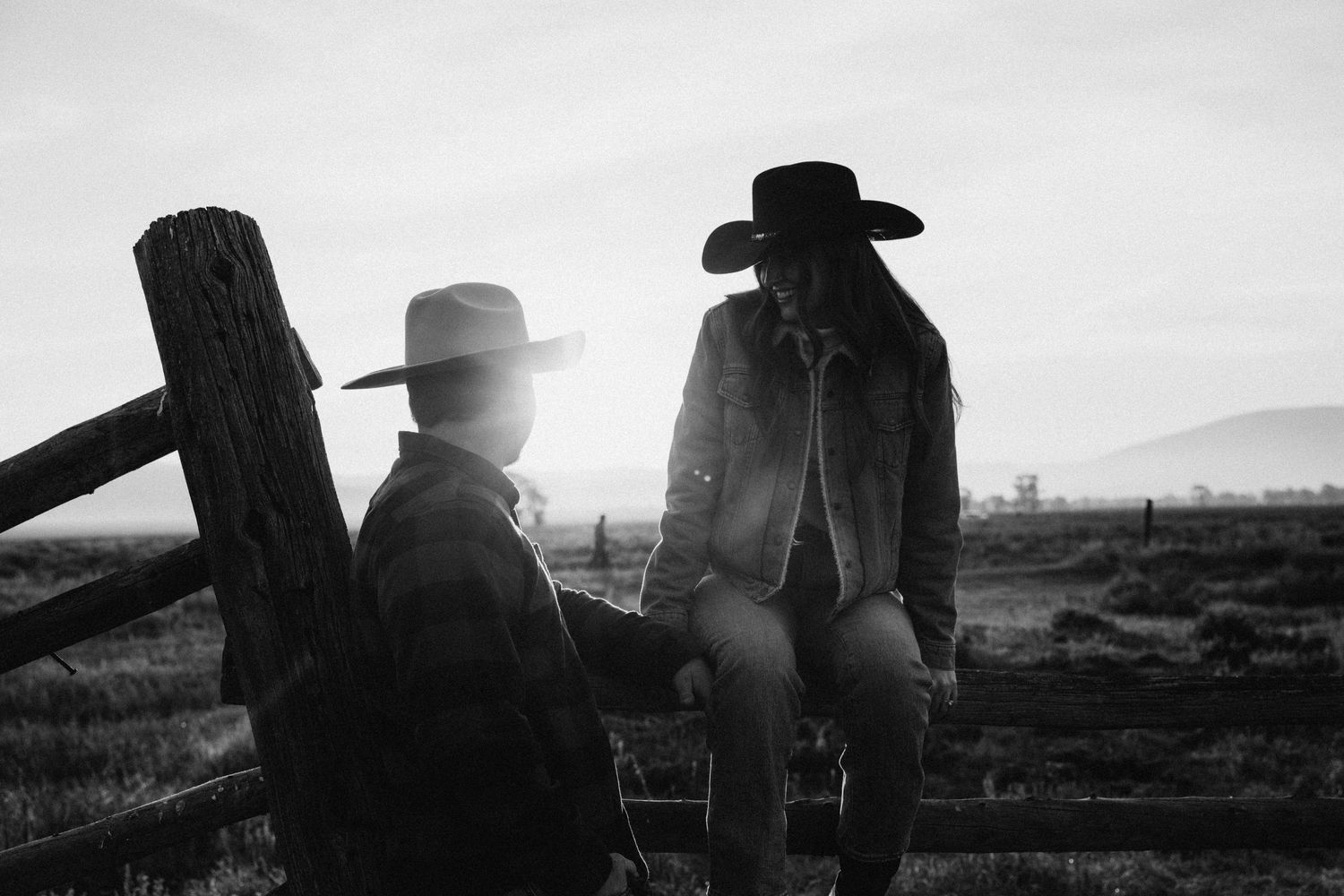 Two figures in cowboy hats sit on a fence silhouetted against a misty landscape at dusk.