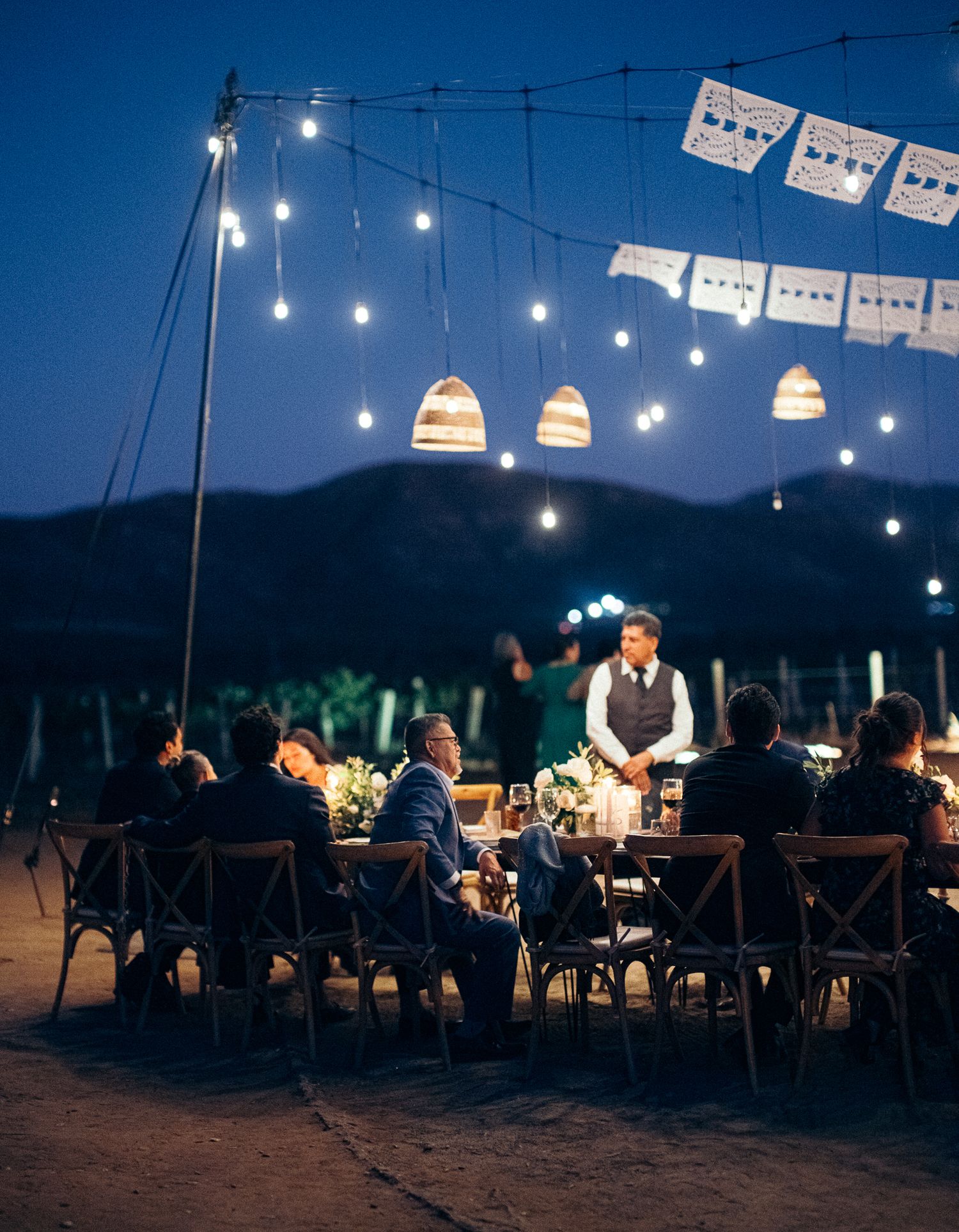 String lights illuminate an outdoor wedding dinner reception against a dusky mountain backdrop.