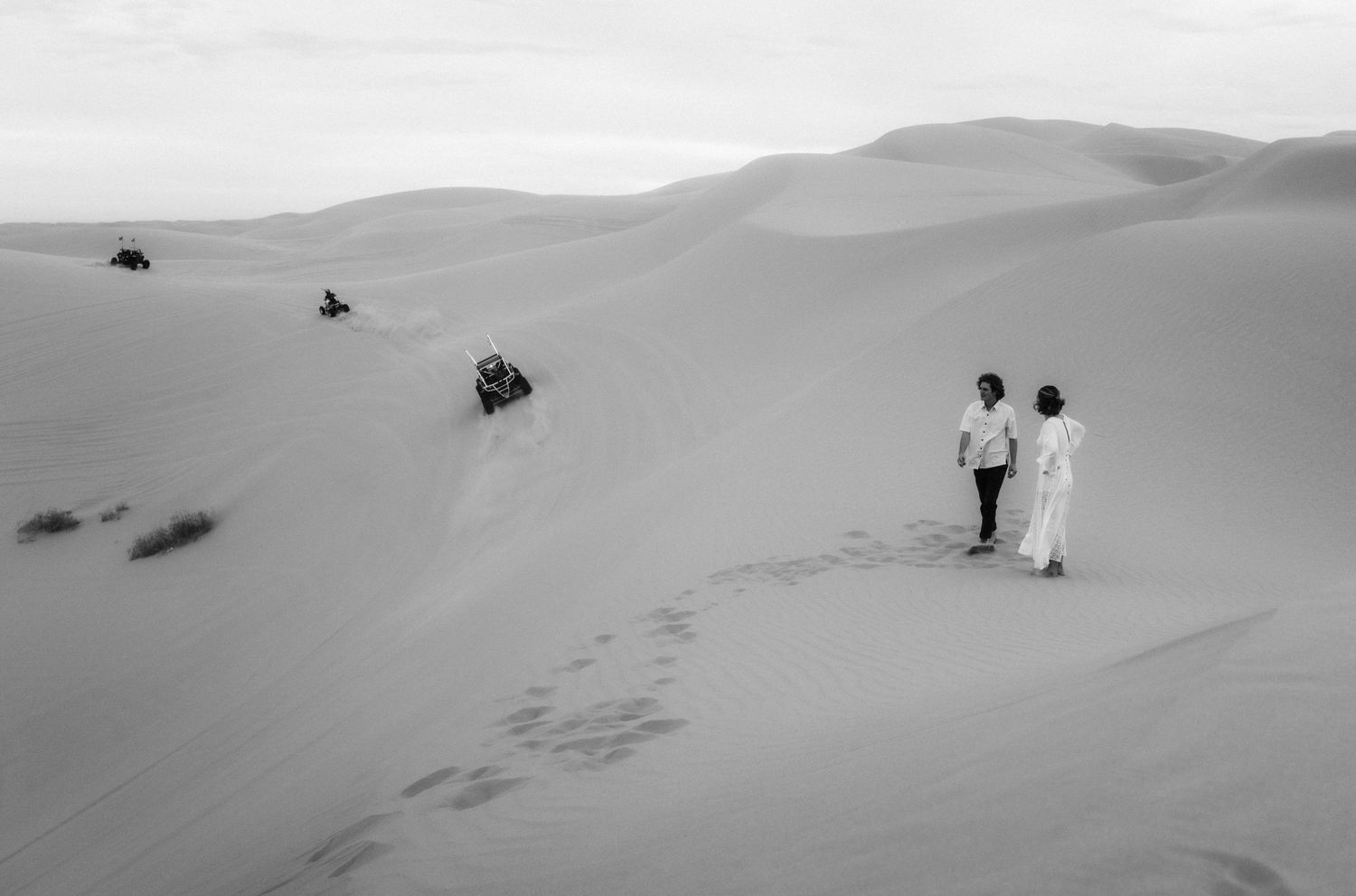 Black and white photograph of figures walking among vast desert sand dunes.
