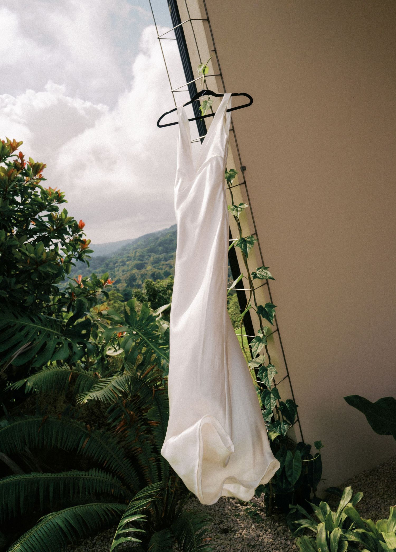 A white wedding dress hanging on a clothes hanger outdoors against lush tropical foliage and mountains in the background.
