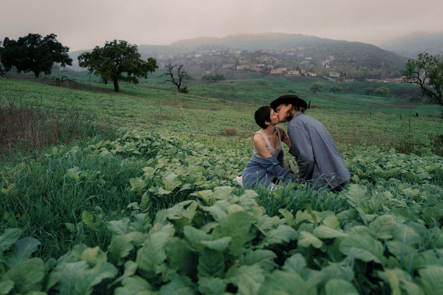 Two people share an intimate moment in a lush green hillside garden at dusk.