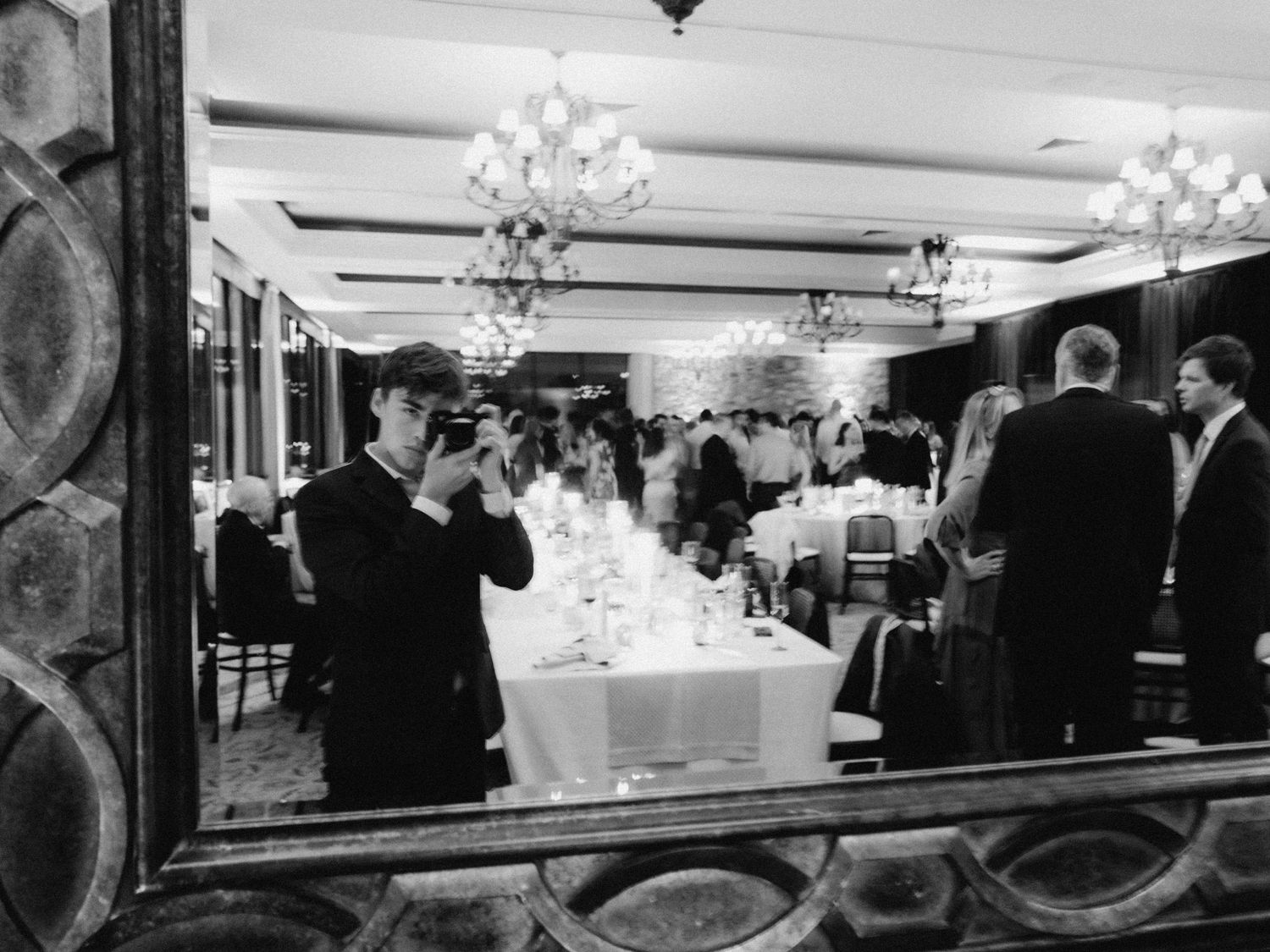 Black and white view of an elegant restaurant dining room with crystal chandeliers and people seated at formal tables.