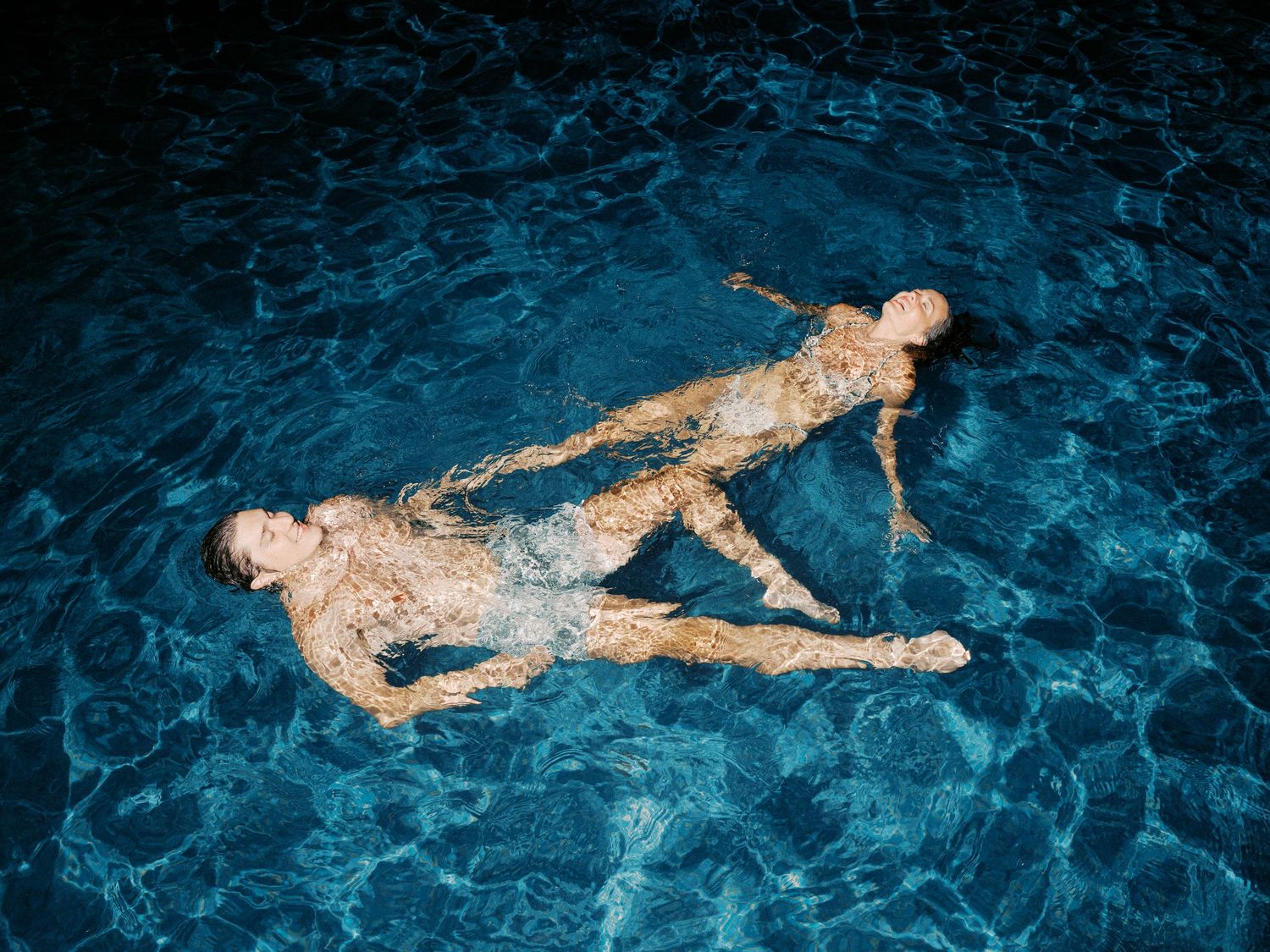 Two swimmers float peacefully on bright turquoise water in a night swimming scene illuminated from below.