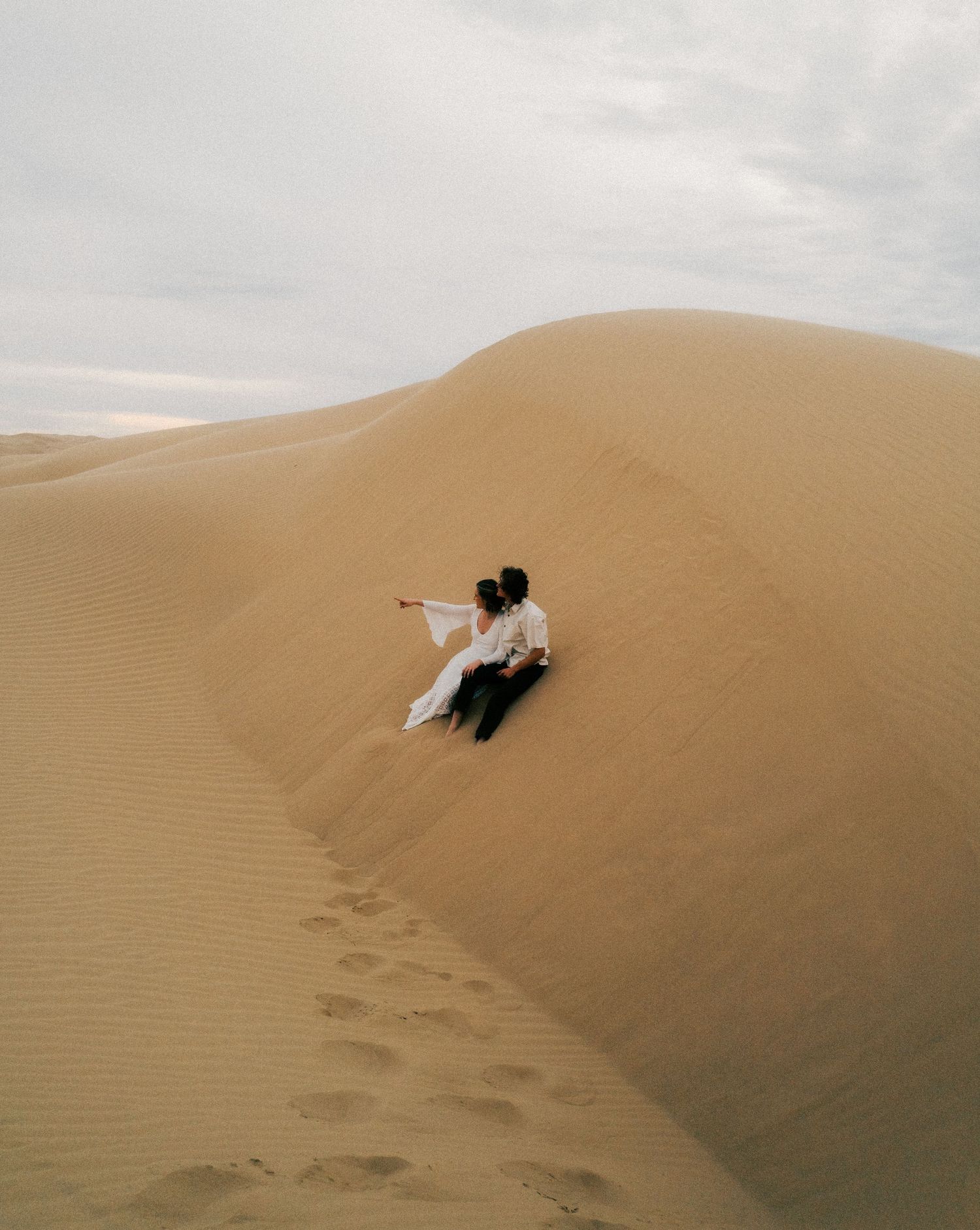 A person lays on the edge of a sand dune in a desert landscape under cloudy skies with footprints visible below.