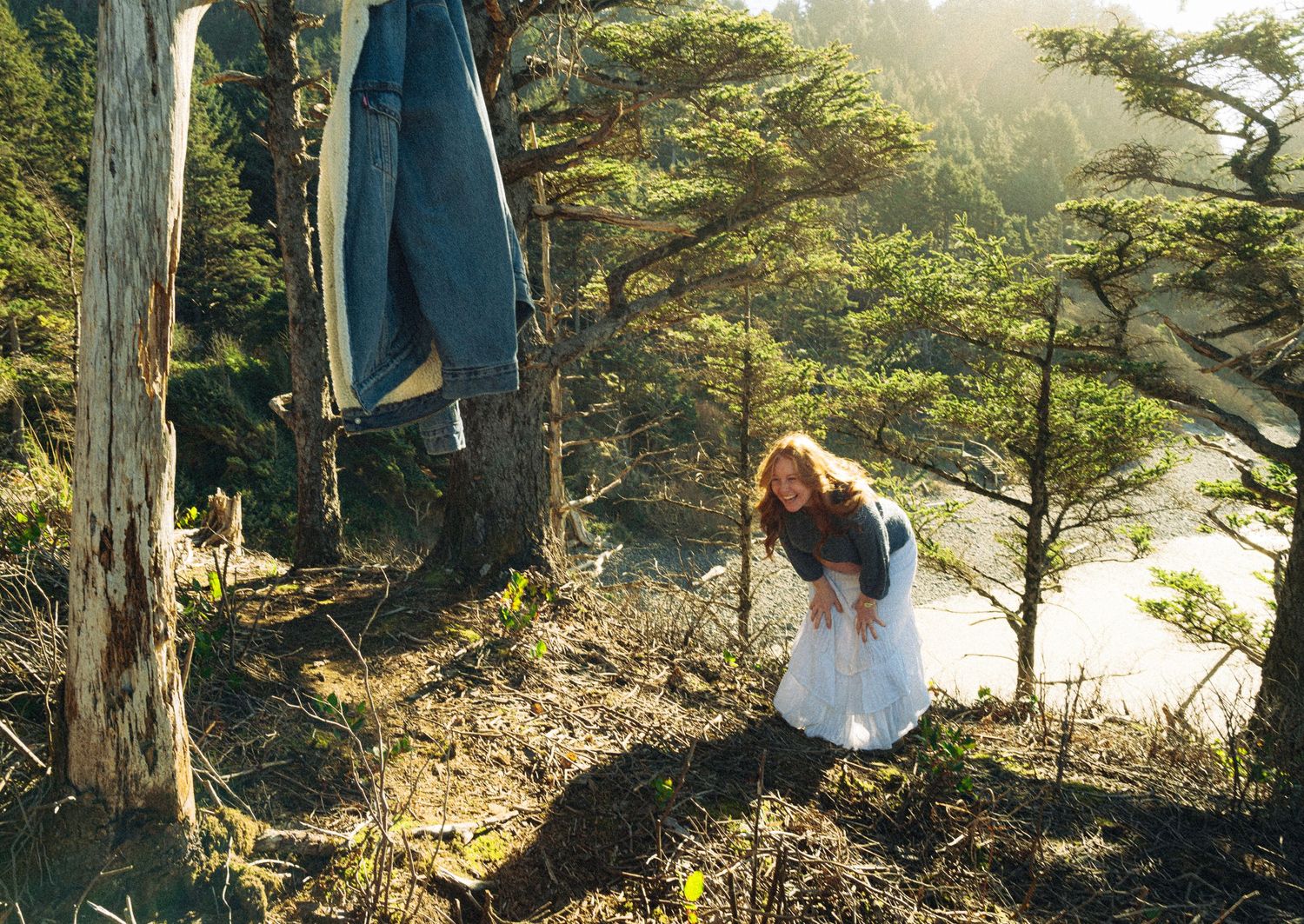 White dress flows in sunlit forest setting among tall trees.