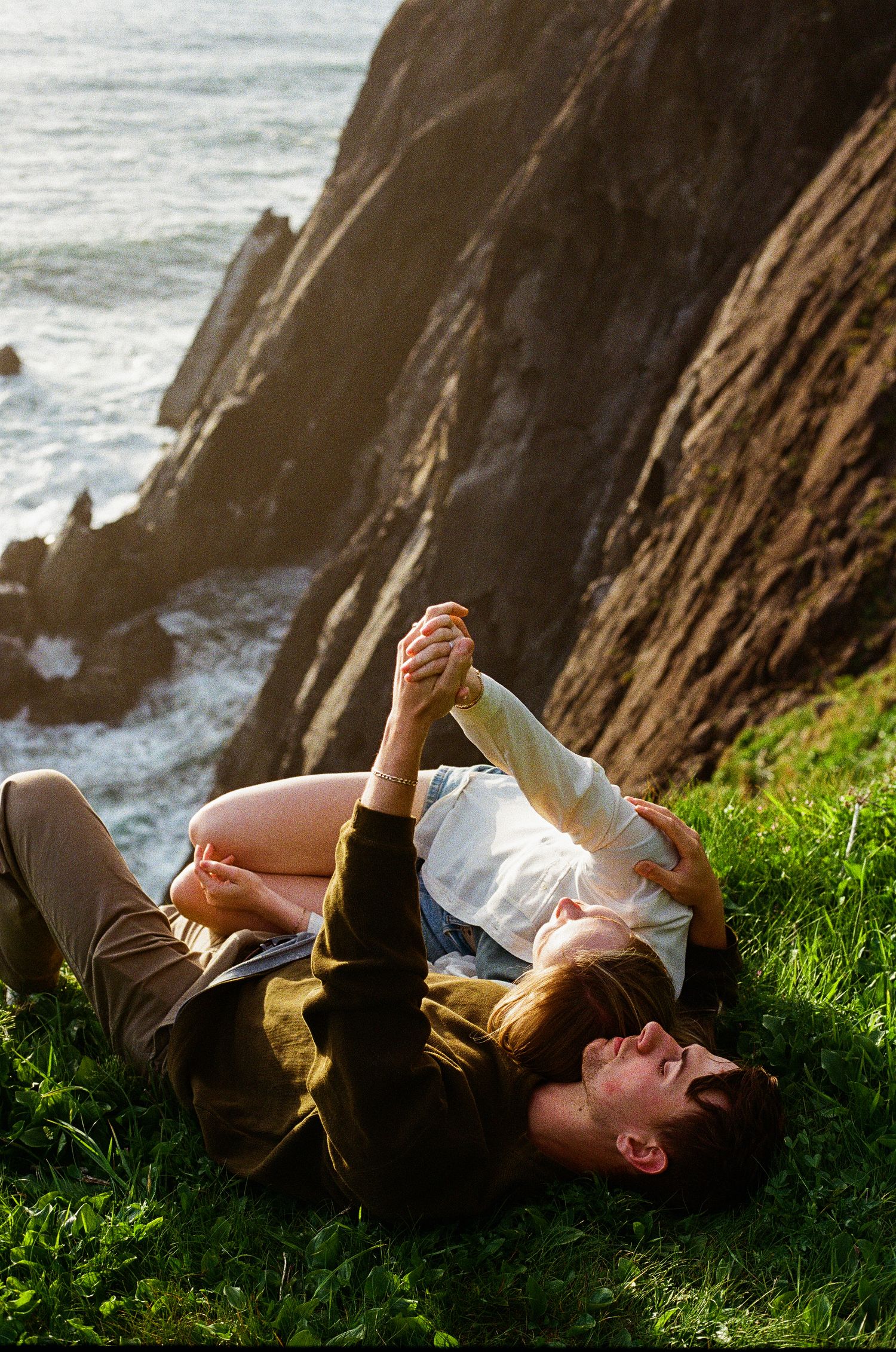 A couple relaxes on grassy cliffs overlooking dramatic ocean waves and rocky coastline.