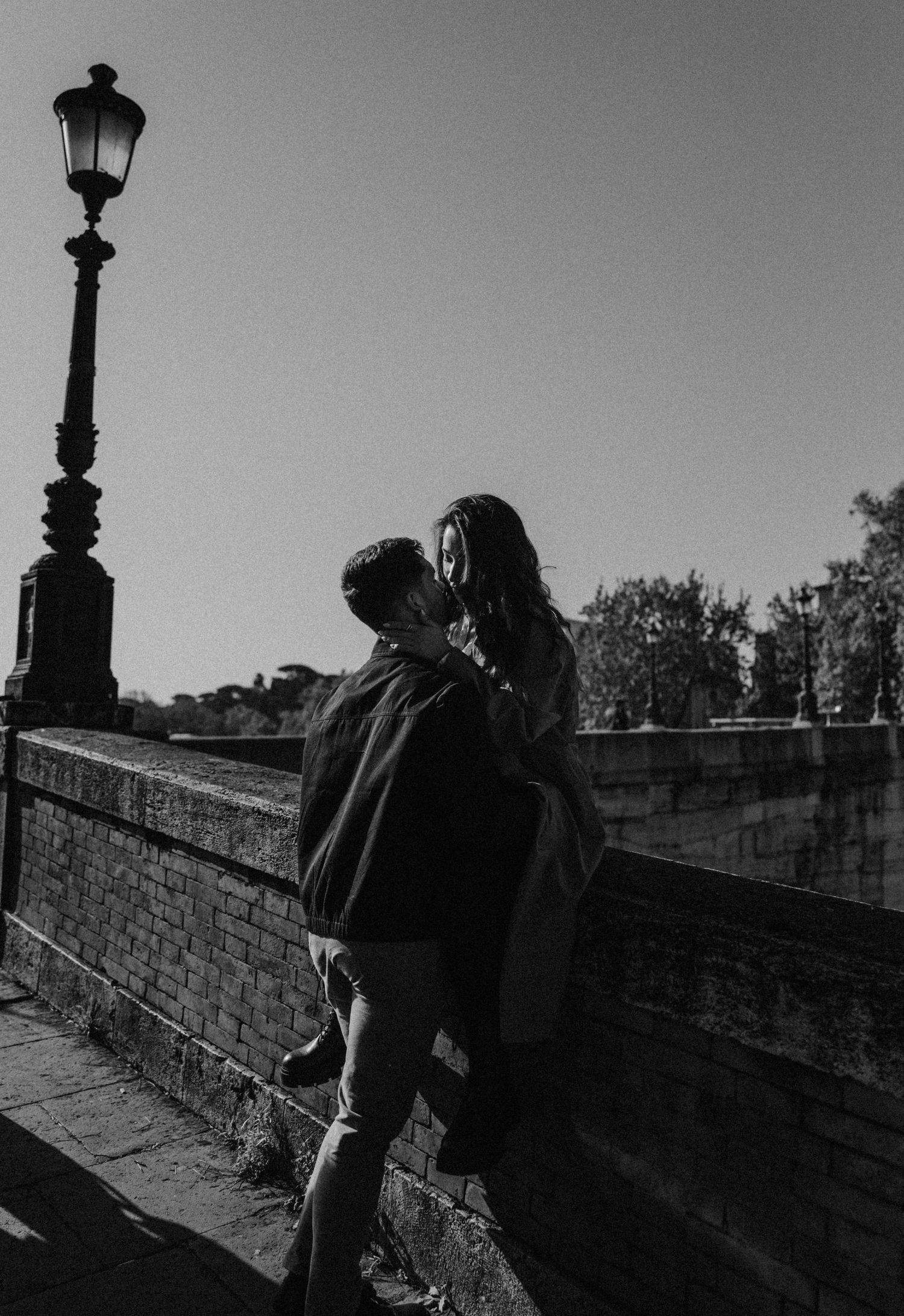 A romantic black and white photograph of a couple embracing near a vintage streetlamp on a brick wall.