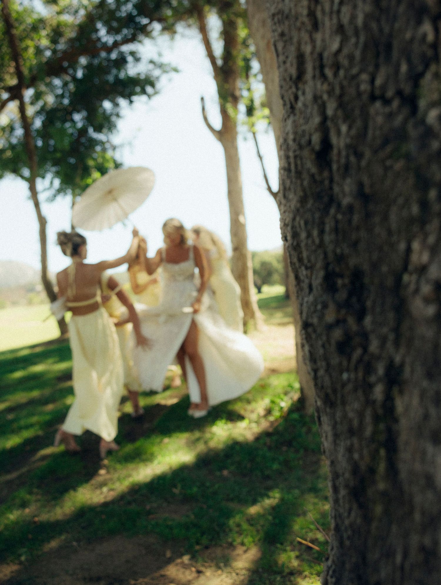 Playful wedding party members with parasols dance in dappled sunlight under trees.