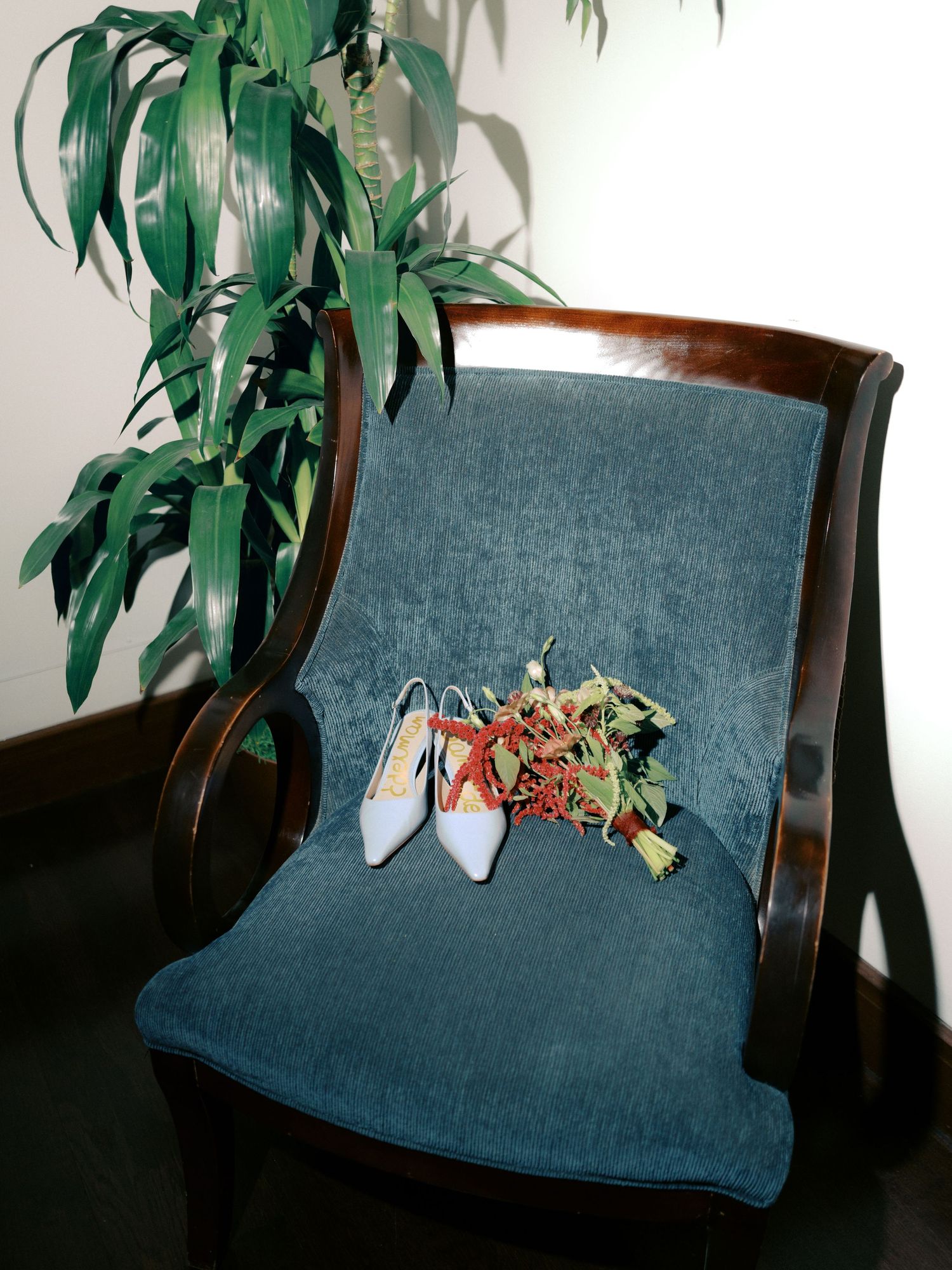 Orange ballet shoes and dried flowers rest on a navy blue upholstered armchair with curved wooden arms near a green plant.