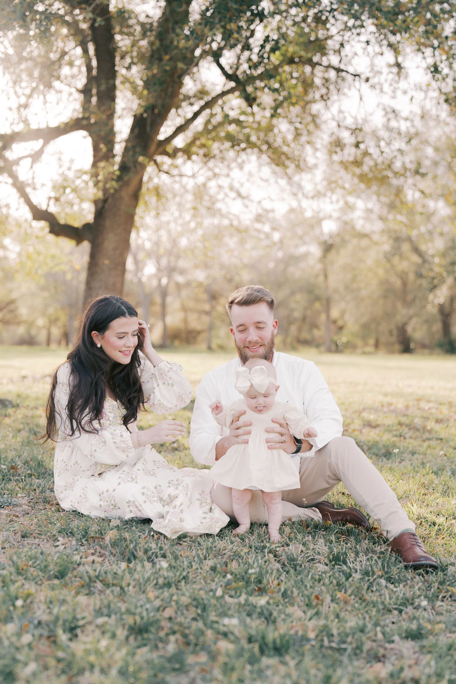 Family in white outfits enjoying a tender moment on grass under a large tree.
