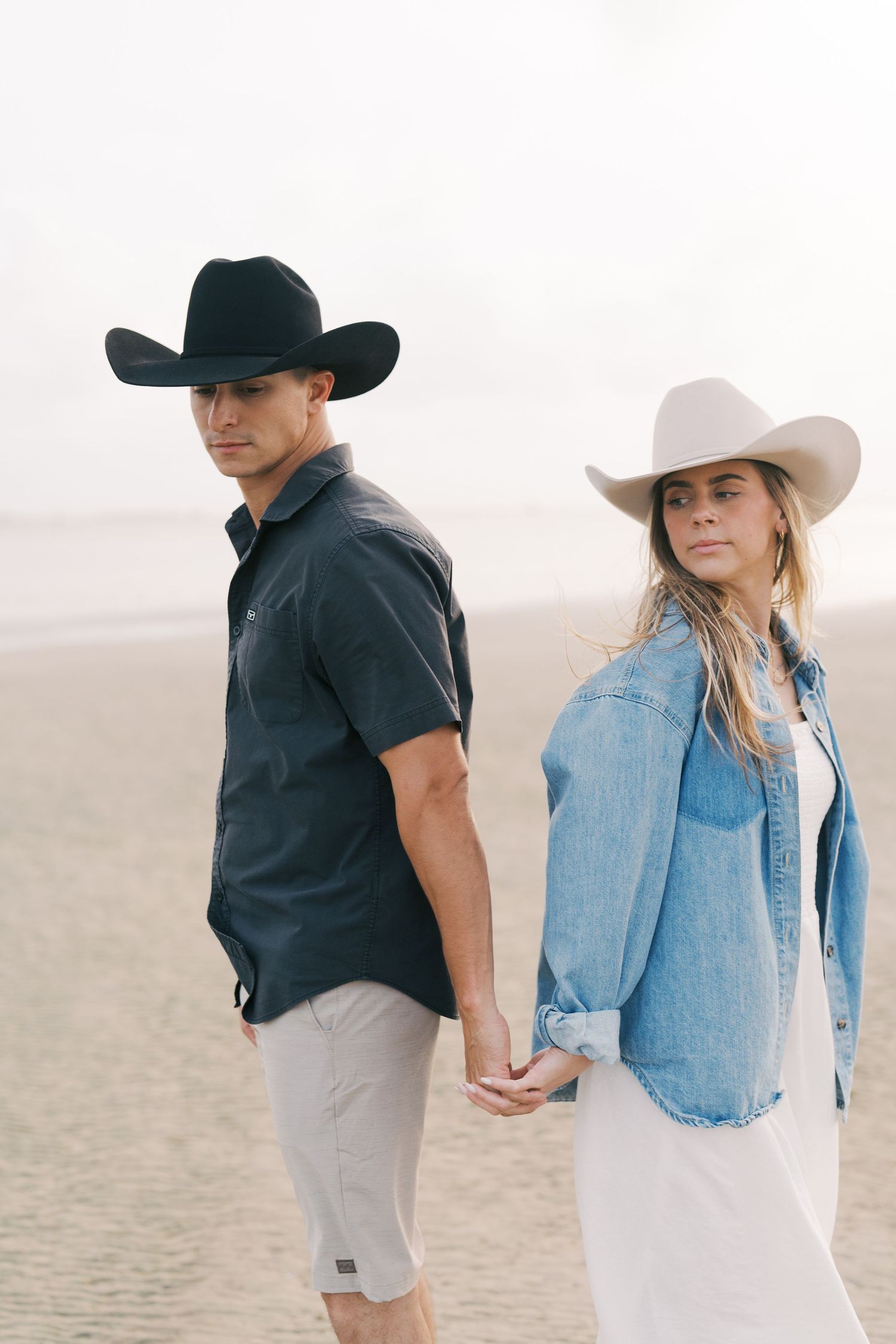 A couple in western attire holding hands on a beach while wearing cowboy hats.