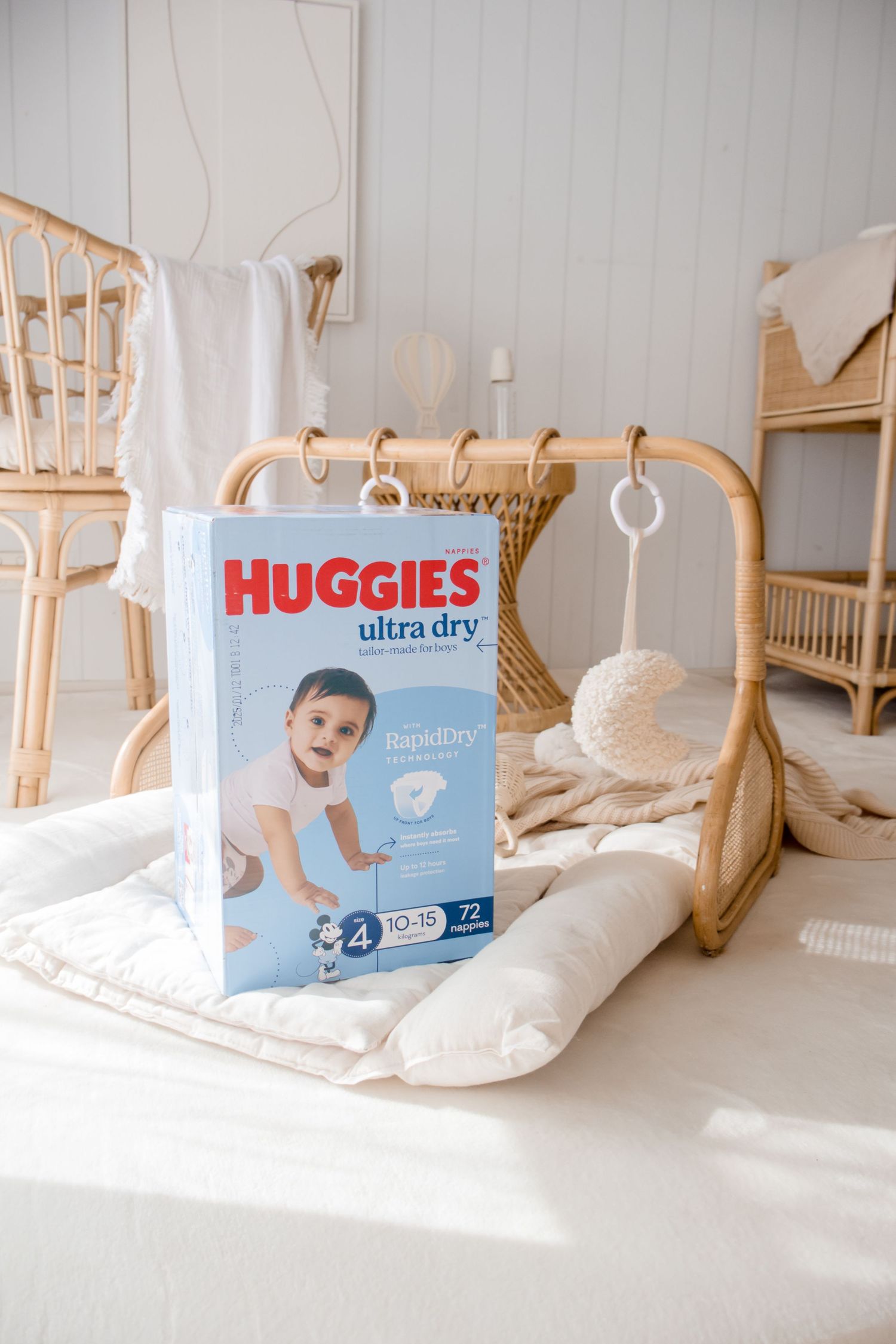 A box of Huggies Ultra Dry diapers sits on a white surface with rattan furniture and baby accessories in the background.